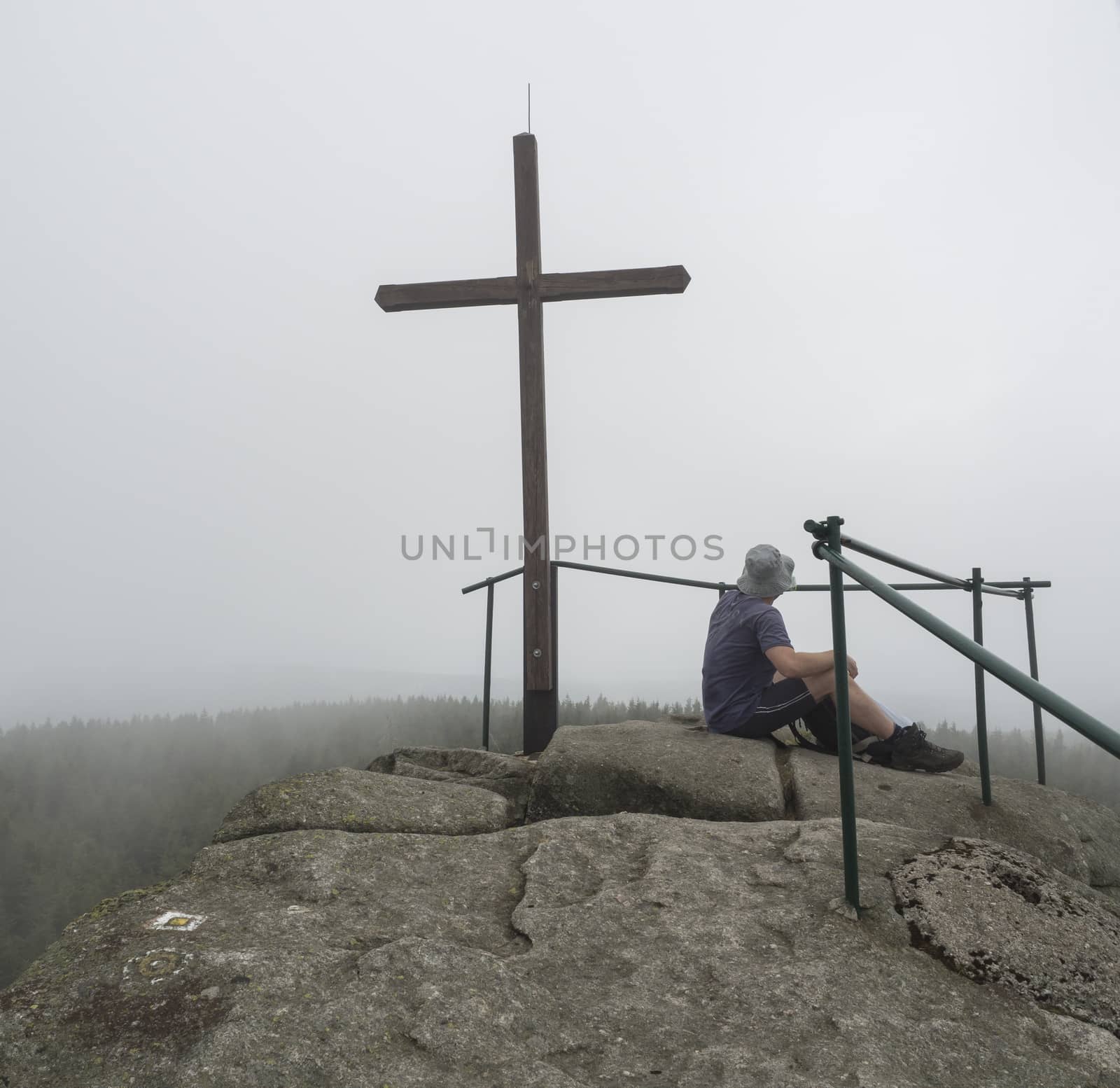 young hiker man sitting on the top peak of Jizera mountain rock with big cross in moody day, fog tree background