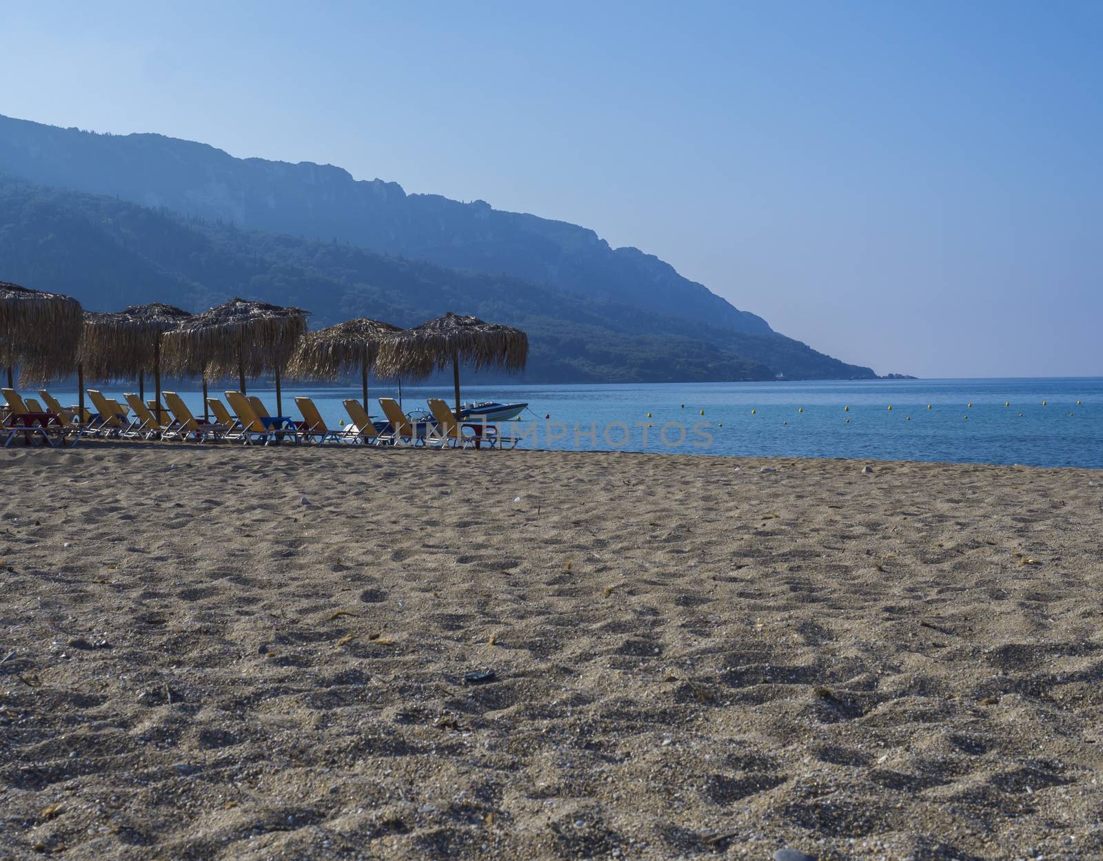 Sand and pebbles beach and turquoise sea shore at Agios Georgios Pagon at Corfu island, Greece with sun umbrellas orange sunbeds and boat, green hill and clear blue sky backgound.