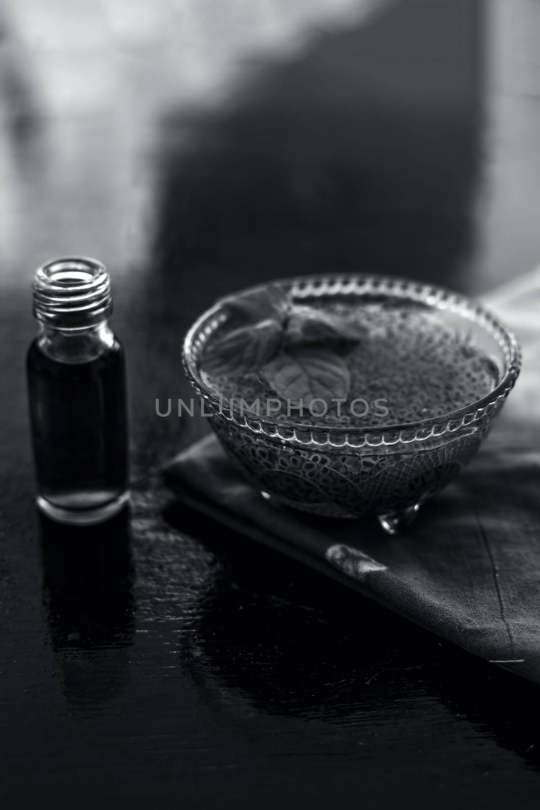 Glass bowl full of soaked sabja seeds or falooda seeds or sweet basil seeds with its extracted essence or essential oil in a transparent glass bottle on wooden surface.