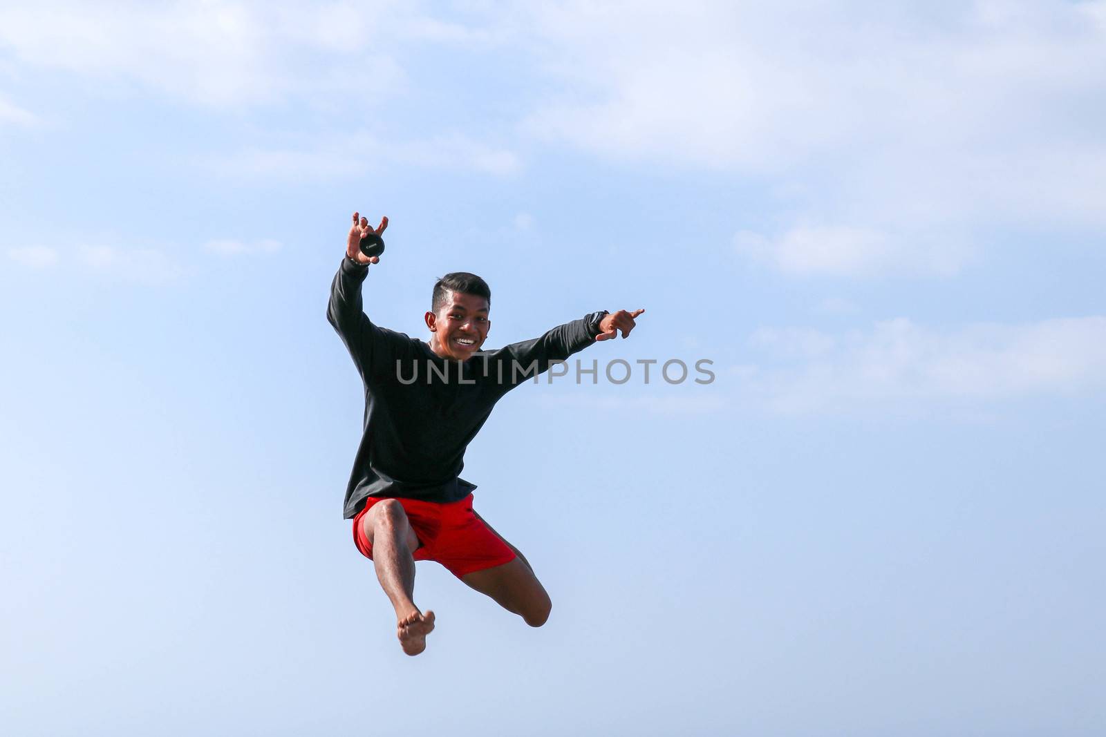 Man jumping happy in the beach with a blue sky in the background. Young cheerful Indonesian jumps. A teenager in a good mood jumps in the air. Background with blue sky. Sunny tropical day.