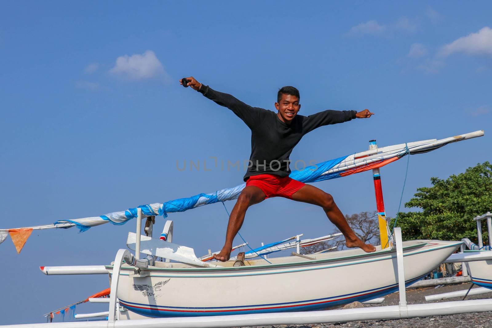 Happy young man jumping on the beach. Young cheerful Indonesian jumps. A teenager in a good mood jumps in the air. In the background a fishing boat. Sunny tropical day with blue sky.