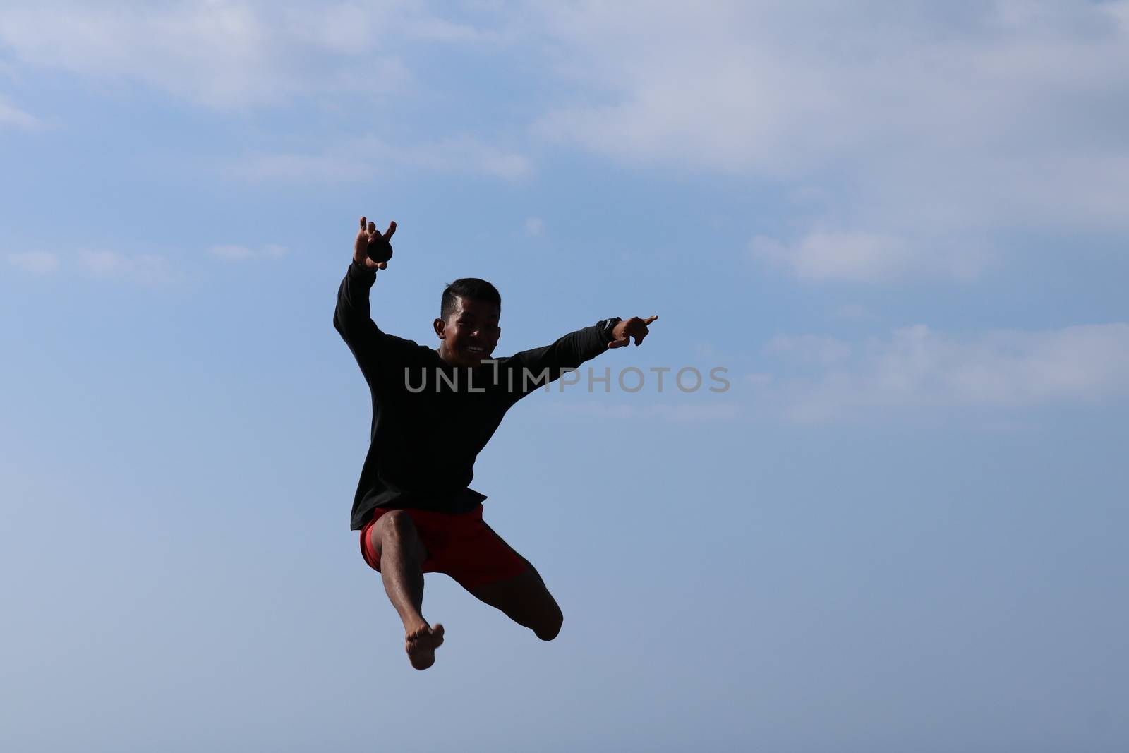 Happy han jumping above blue sky background. Young cheerful Indonesian jumps. A teenager in a good mood jumps in the air. Sunny tropical day.