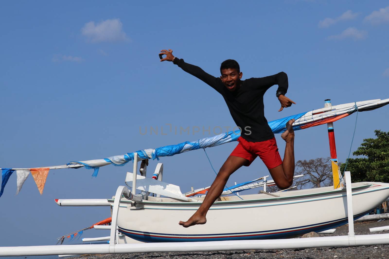 Happy young Indonesian man jumps on the beach. A teenager in a good mood jumps in the air. Sunny tropical day with blue sky.
