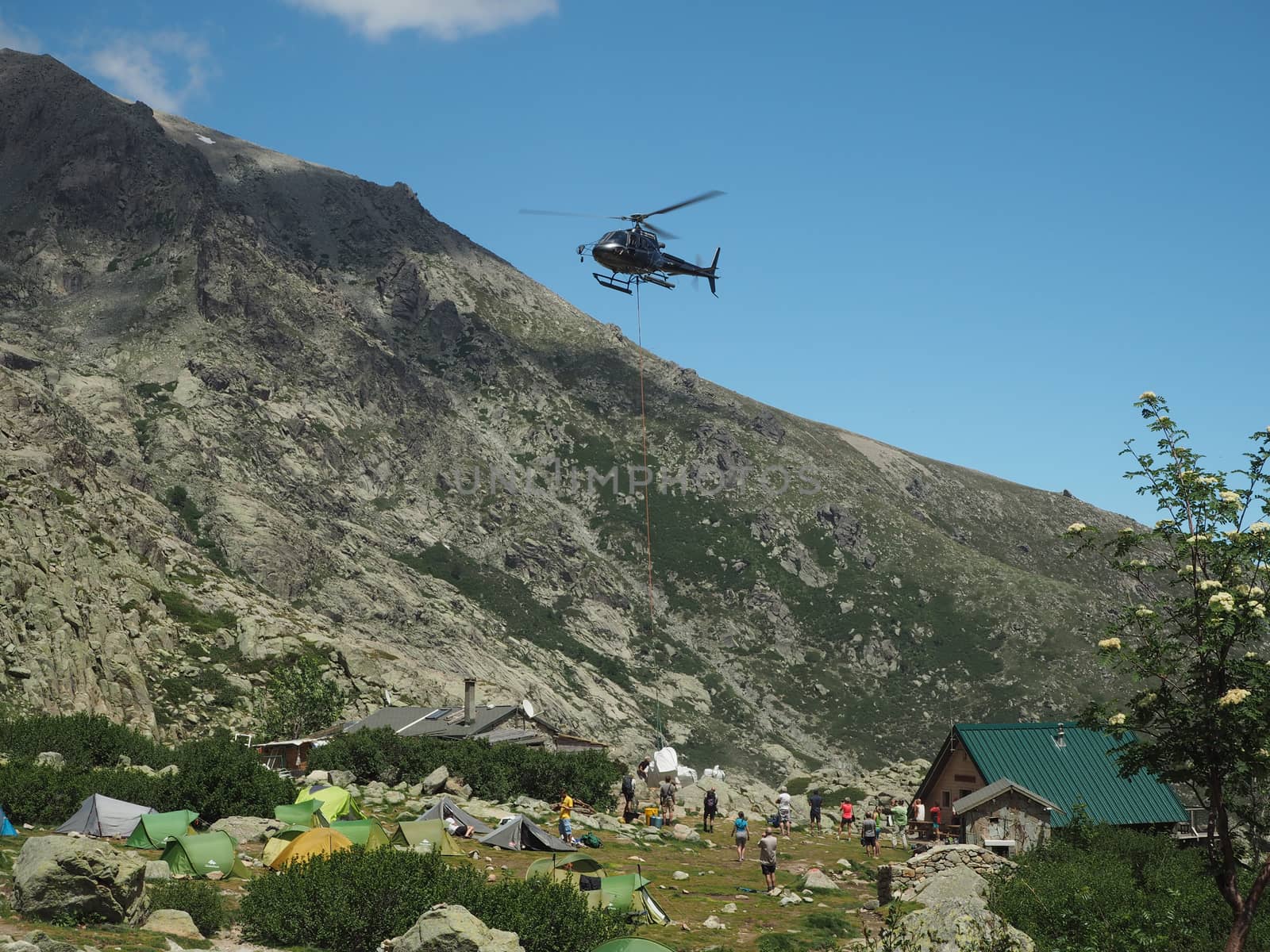 France, Corsica, Corsician Alps, June 19, 2017: helicopter dropping off supplies for mountain camp Refuge de Pietra Piana on famous hiking trail GR20, rocks, tents, tree and blue sky background by Henkeova