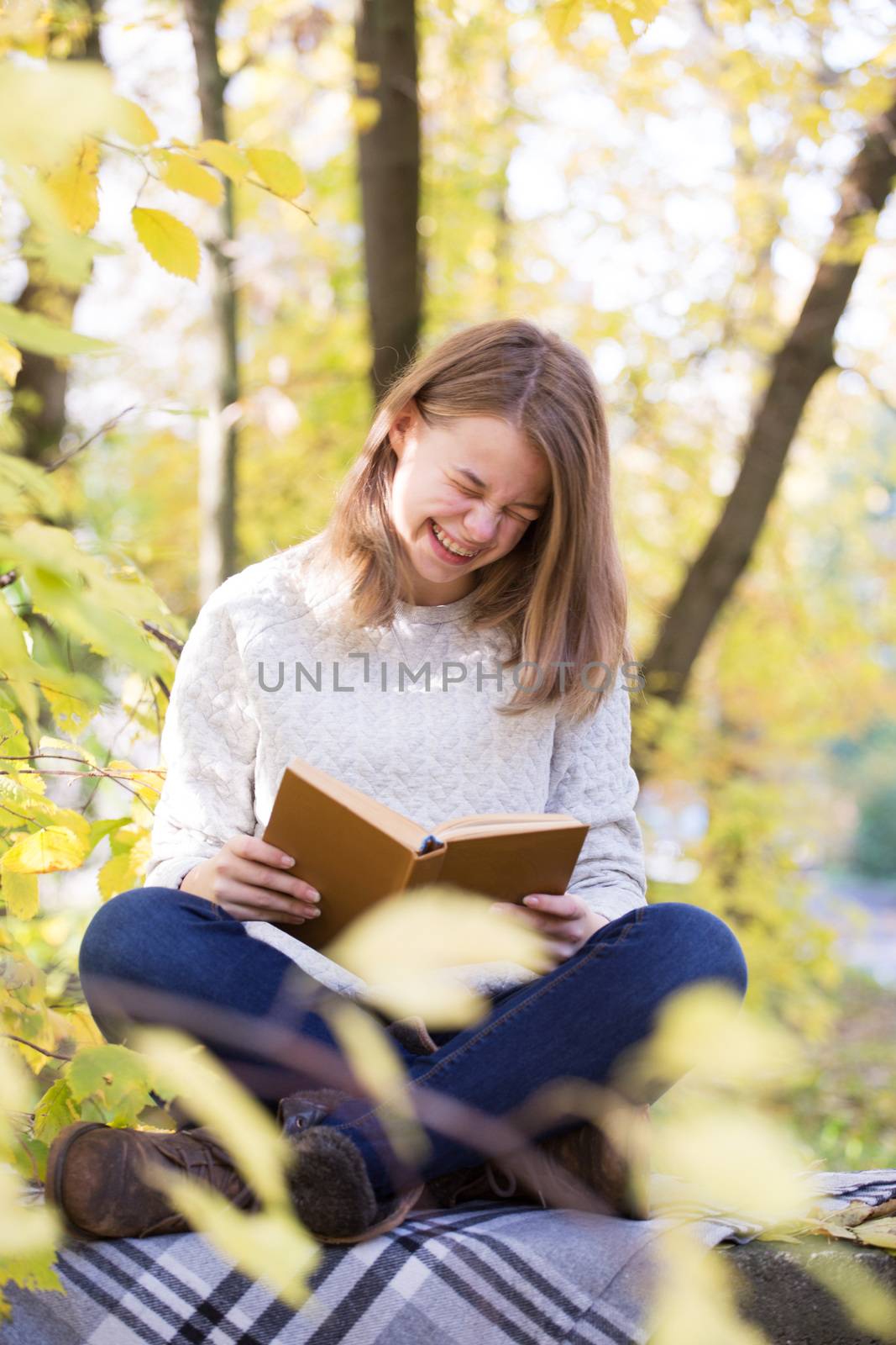 Beautiful teenager girl sitting in autumn garden on the fence , on woolen plaid blanket and laughing reading funny book, dental braces