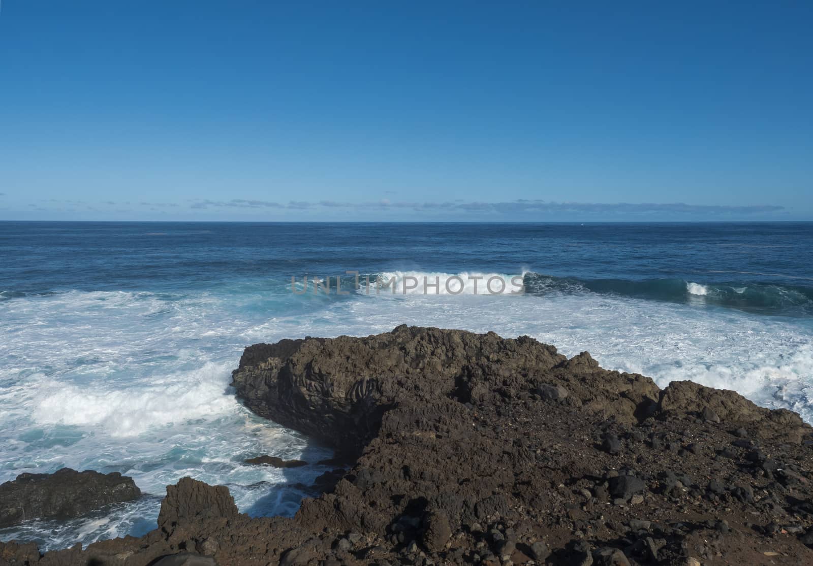 Volcanic rock formation, cliffs of black lava on the rocky shore with crushing white waves over the Atlantic Ocean. Blue sky background. La Palma, Canary Islands