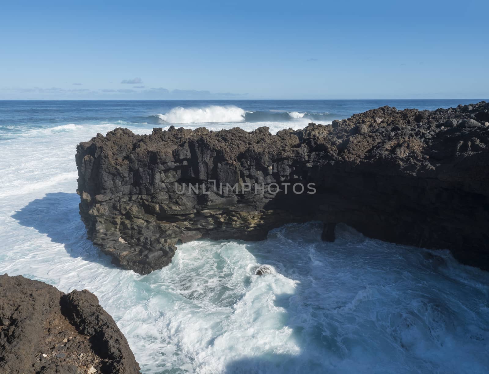 Volcanic rock formation, cliffs of black lava on the rocky shore with crushing white waves over the Atlantic Ocean. Blue sky background. La Palma, Canary Islands