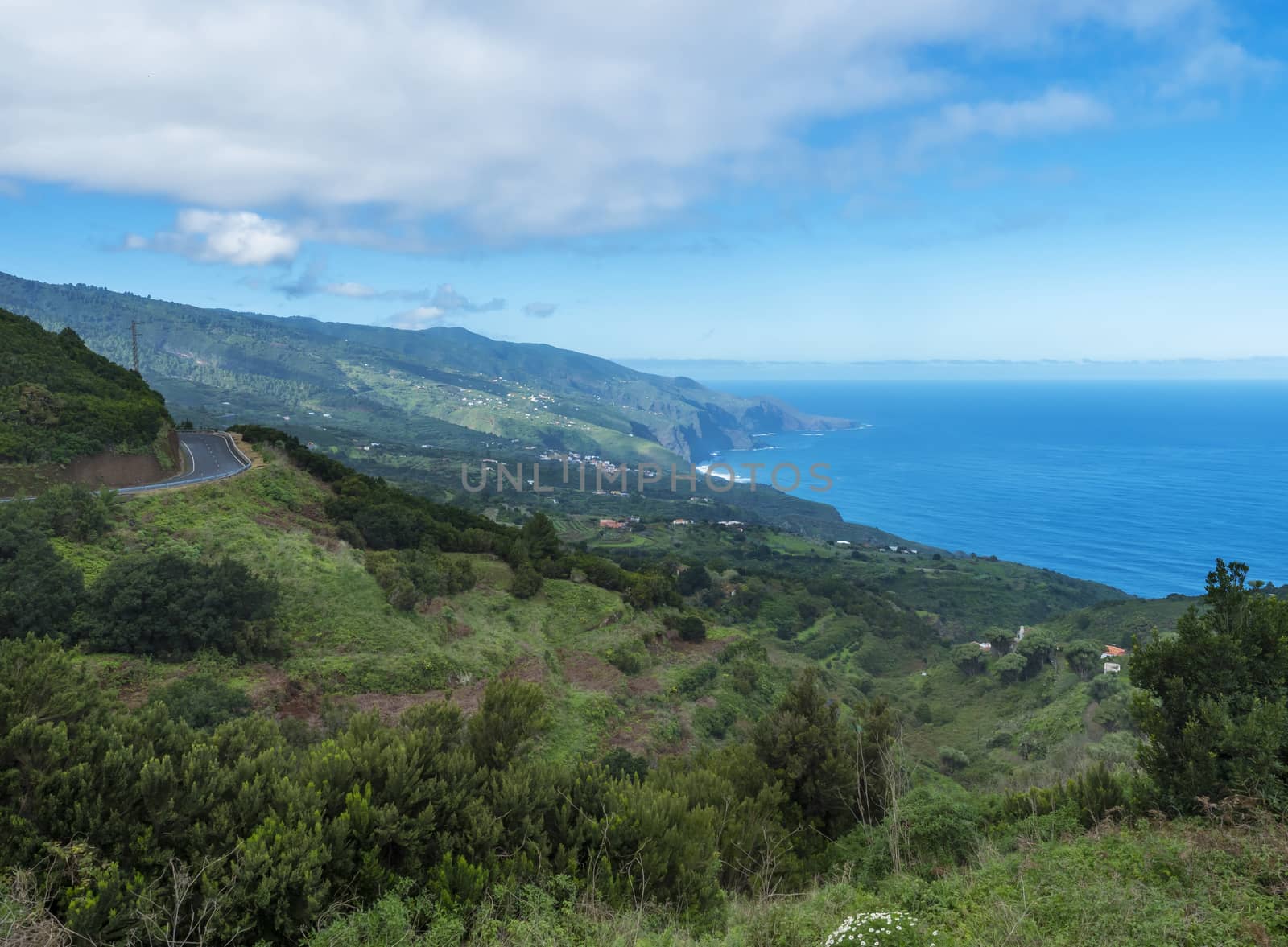 Subtropical landscape with green hills, tree, houses, winding alphalt road and sea horizon. Blue sky white clouds background, La Palma, Canary Islands, Spain.
