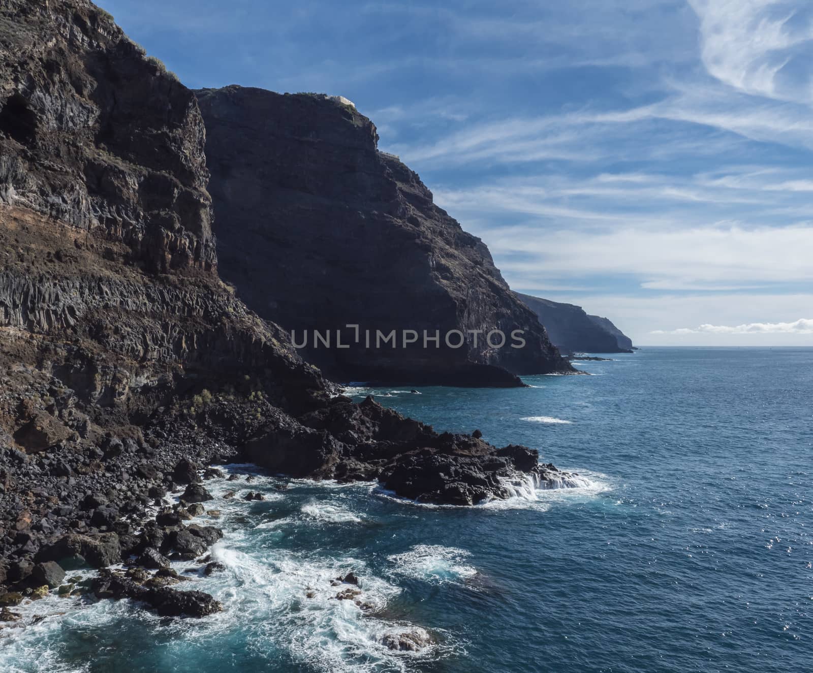 High steep Volcanic rock cliffs of black lava on the rocky shore with crushing white waves over the Atlantic Ocean. Blue sky background. La Palma, Canary Islands