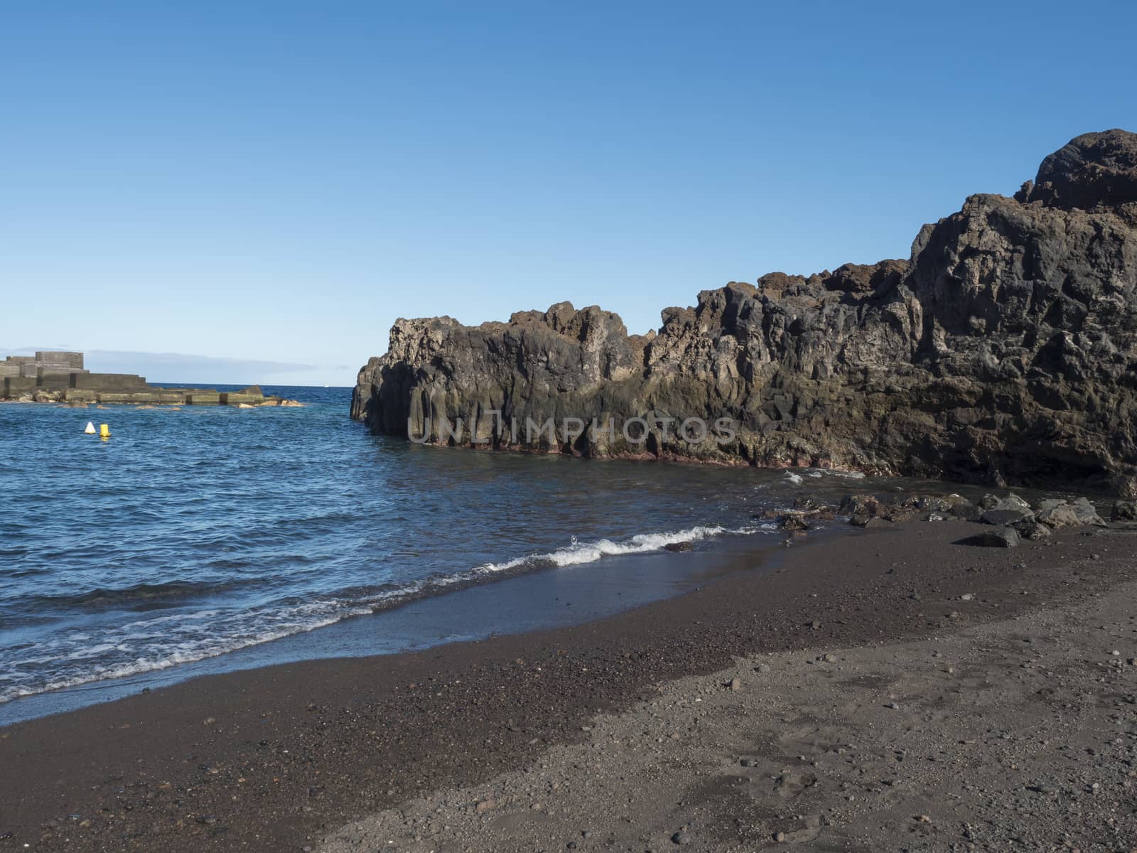 The dark sand beach of the village of Los Cancajos at La Palma, Canary Islands. Lava rock cliffs and Blue sky background