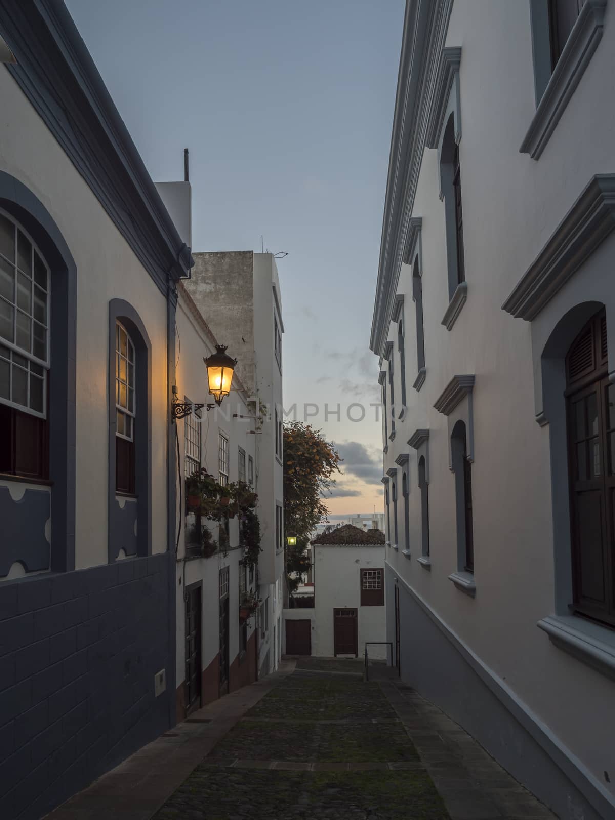 Evening empty narrow street at Santa Cruz de la Palma city center with glowing lantern, blue white traditional houses and cobble stone paving. La Palma, Canary Islands, Spain.