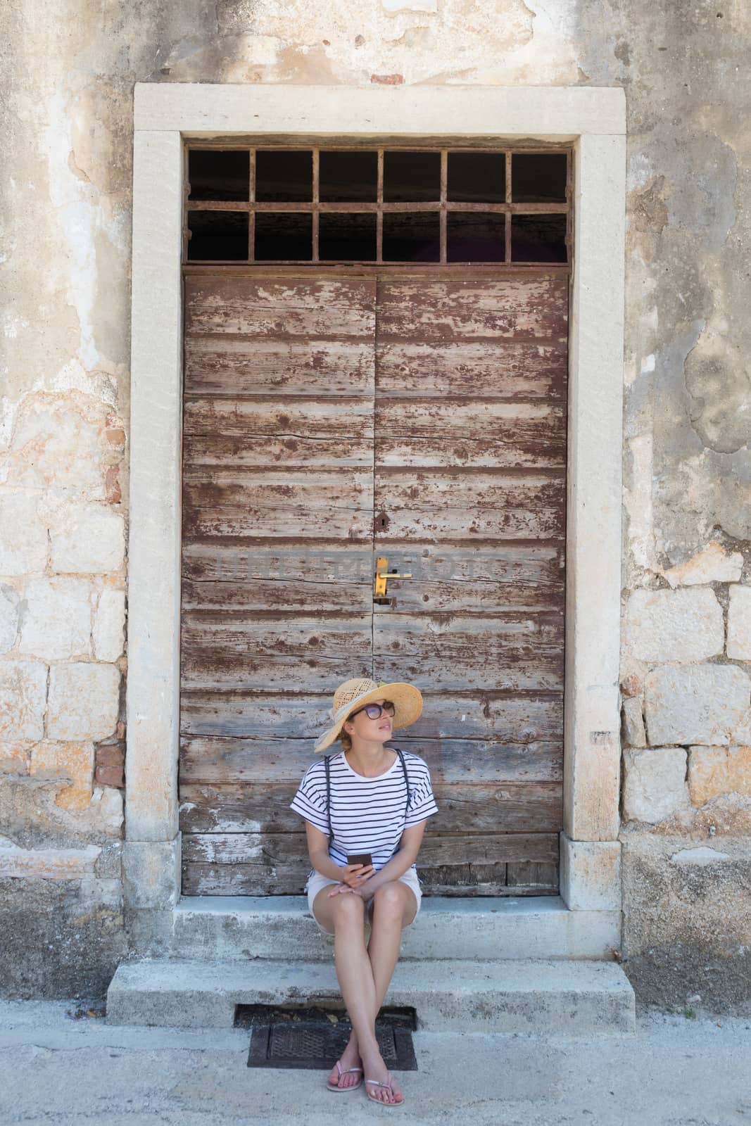 Beautiful young female tourist woman sitting and resting on vinatage wooden doorstep and textured stone wall at old Mediterranean town, smiling, holding, using smart phone to network on vacationes.