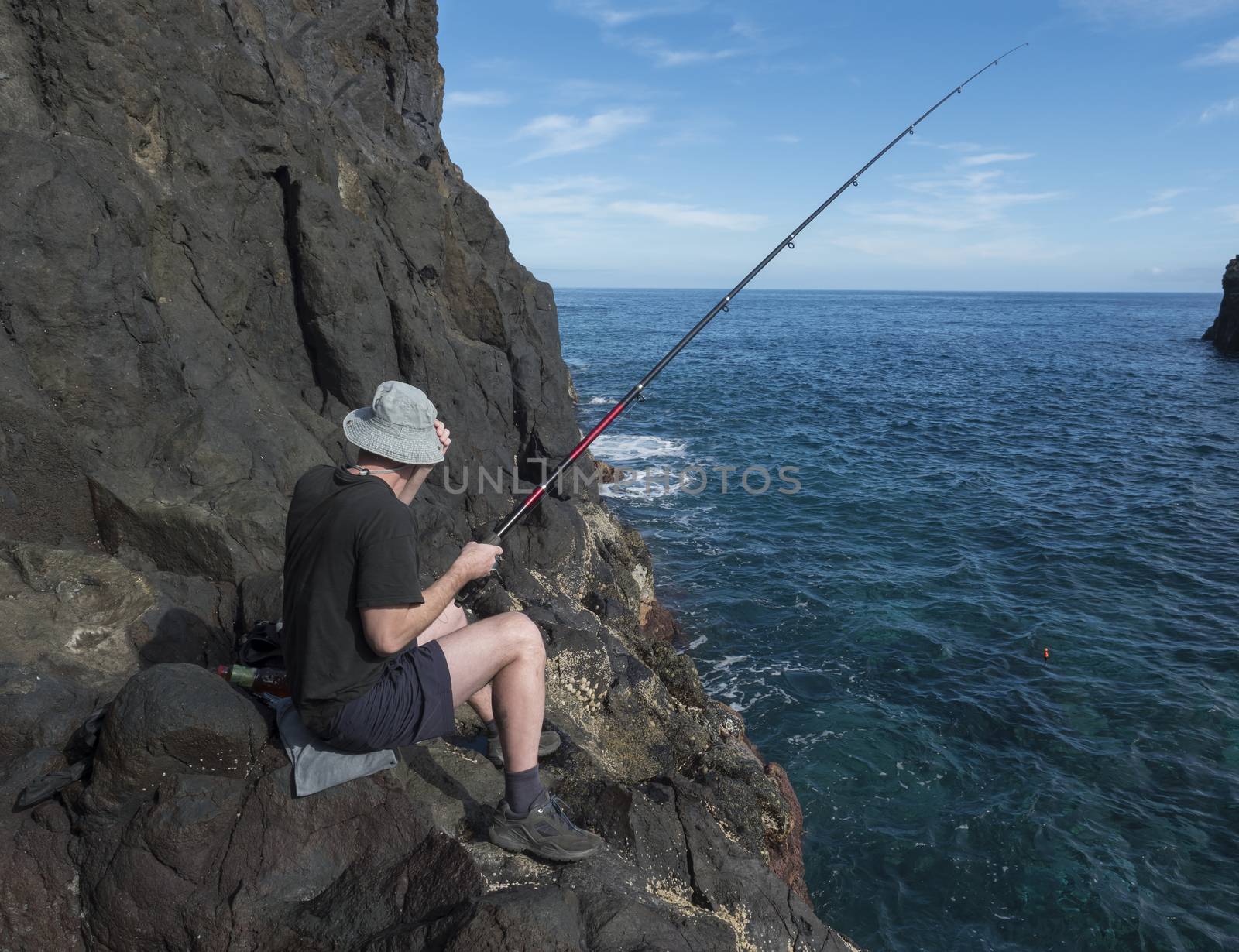 Back side of young man fishermen in hat sitting on the edge of lava rock cliff with fishing rod at coast of atlantic ocean at La Palma island, Spain.