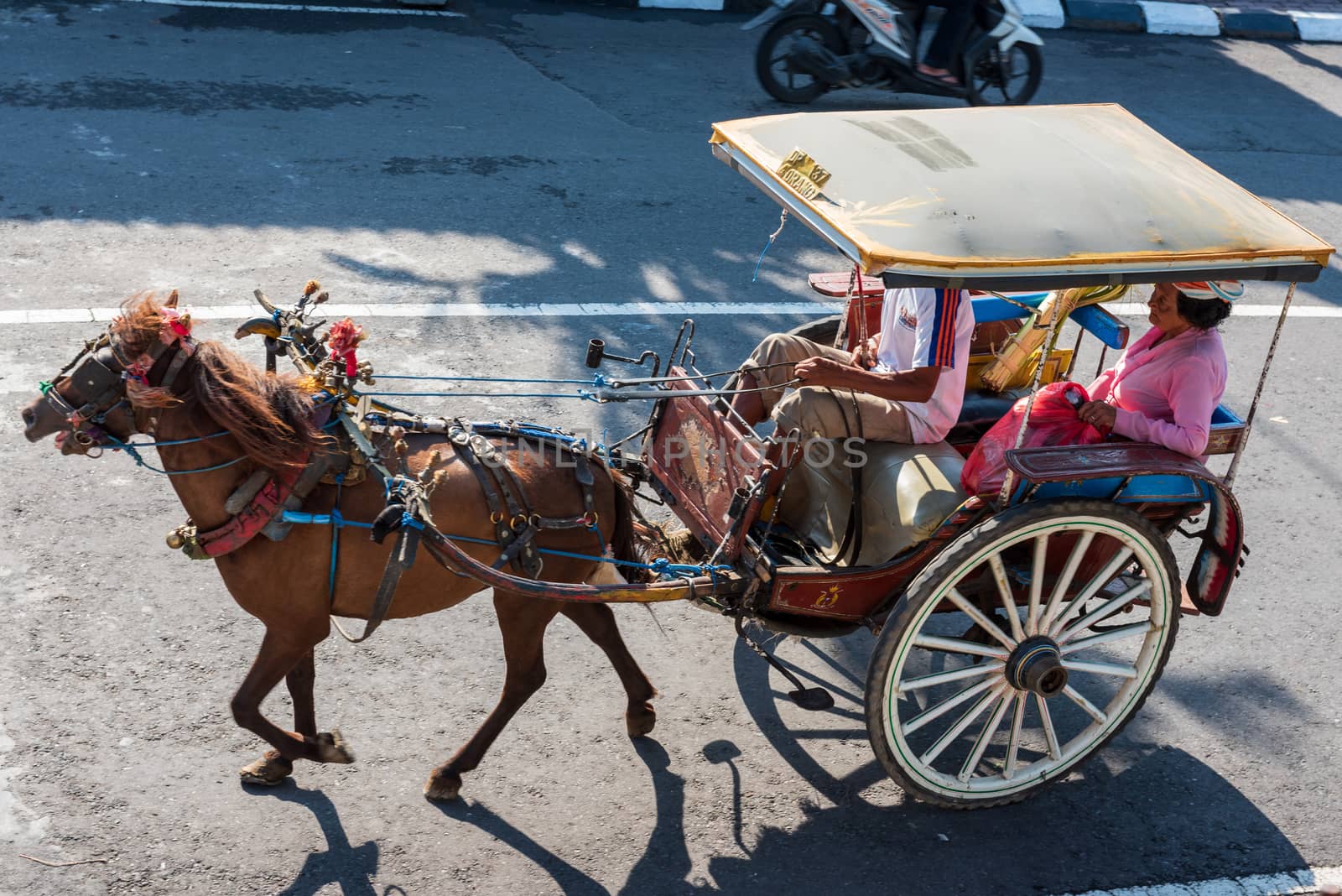 Port Benoa, Bali Indonesia -- Feb 28, 2016. A horse drawn carriage and driver transports a woman passenger wearing a pink outfit and a hat in Bali, In