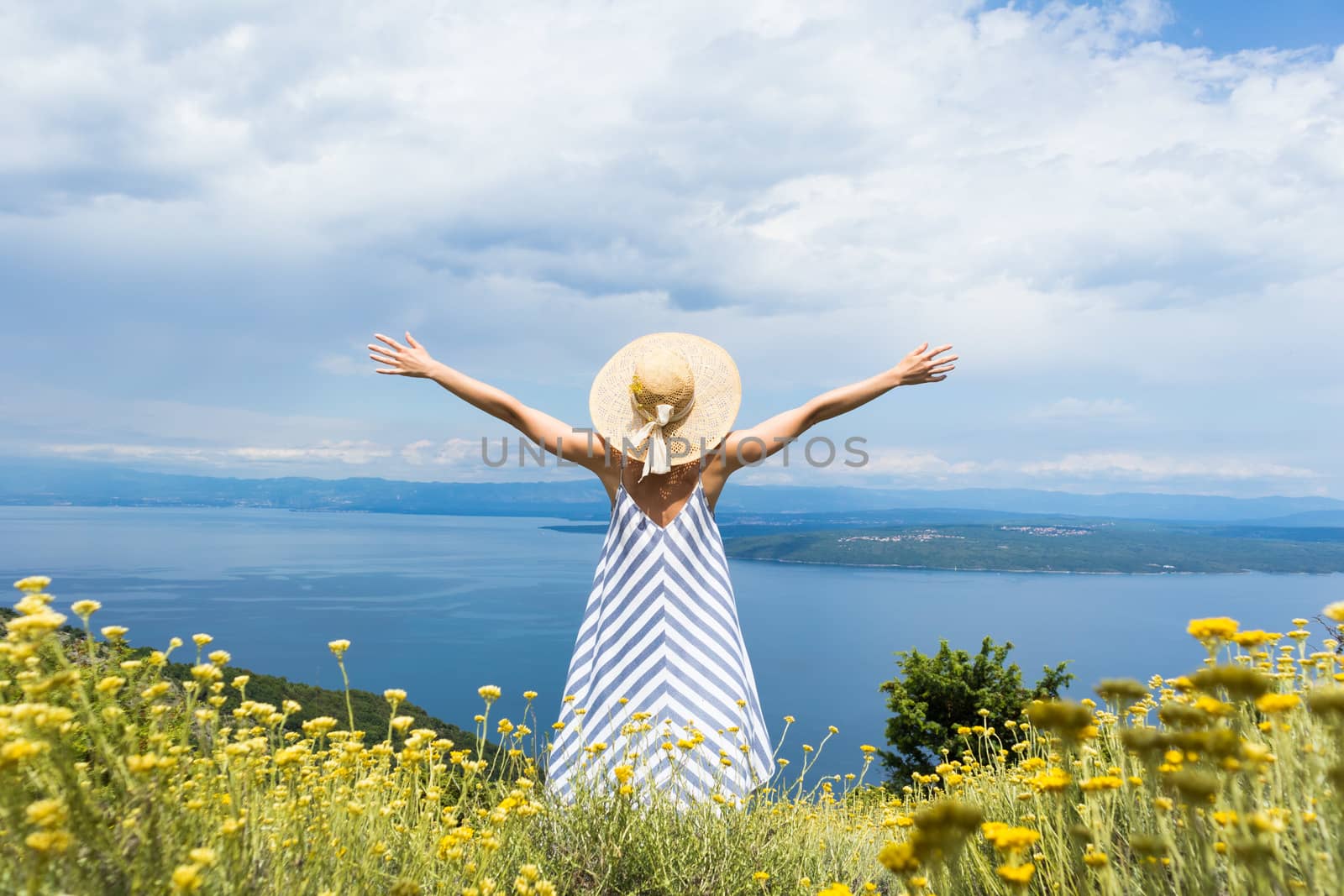 Rear view of young woman wearing striped summer dress and straw hat standing in super bloom of wildflowers, relaxing with hands up to the sky, enjoing beautiful view of Adriatic sea nature, Croatia by kasto