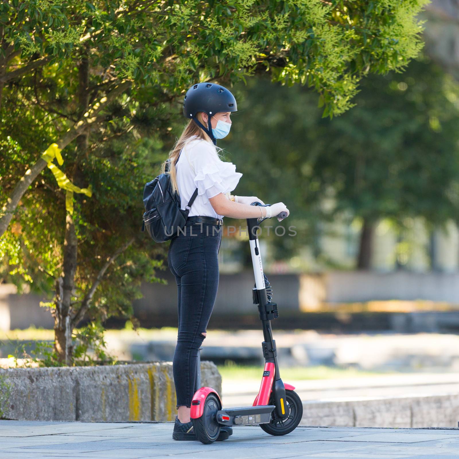 Trendy fashinable teenager girl wearing corona virus protective face mask in public while using rental electric scooters in city environment. New eco-friendly city transport in Ljubljana, Slovenia by kasto