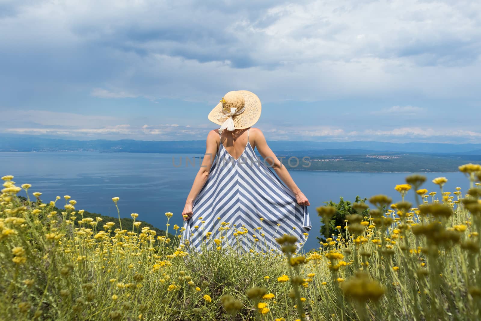 Rear view of young woman wearing striped summer dress and straw hat standing in super bloom of wildflowers, relaxing while enjoing beautiful view of Adriatic sea nature, Croatia by kasto