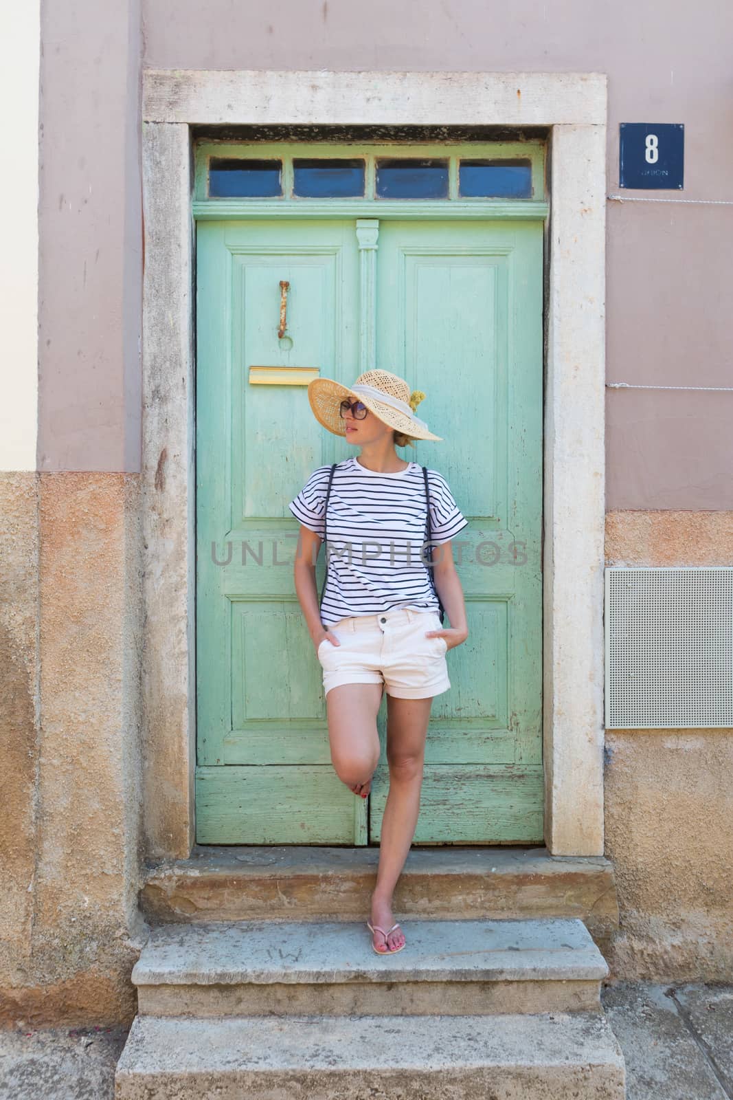 Beautiful young female tourist woman wearing sun hat, standing and relaxing in shade in front of vinatage wooden door in old Mediterranean town while sightseeing on hot summer day by kasto