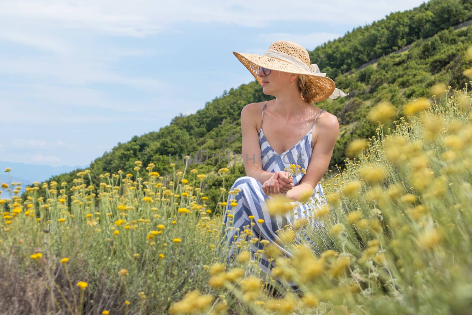 Young woman wearing striped summer dress and straw hat squating in super bloom of wildflowers, relaxing while enjoing beautiful nature of Adriatic sea coastal nature of Croatia.