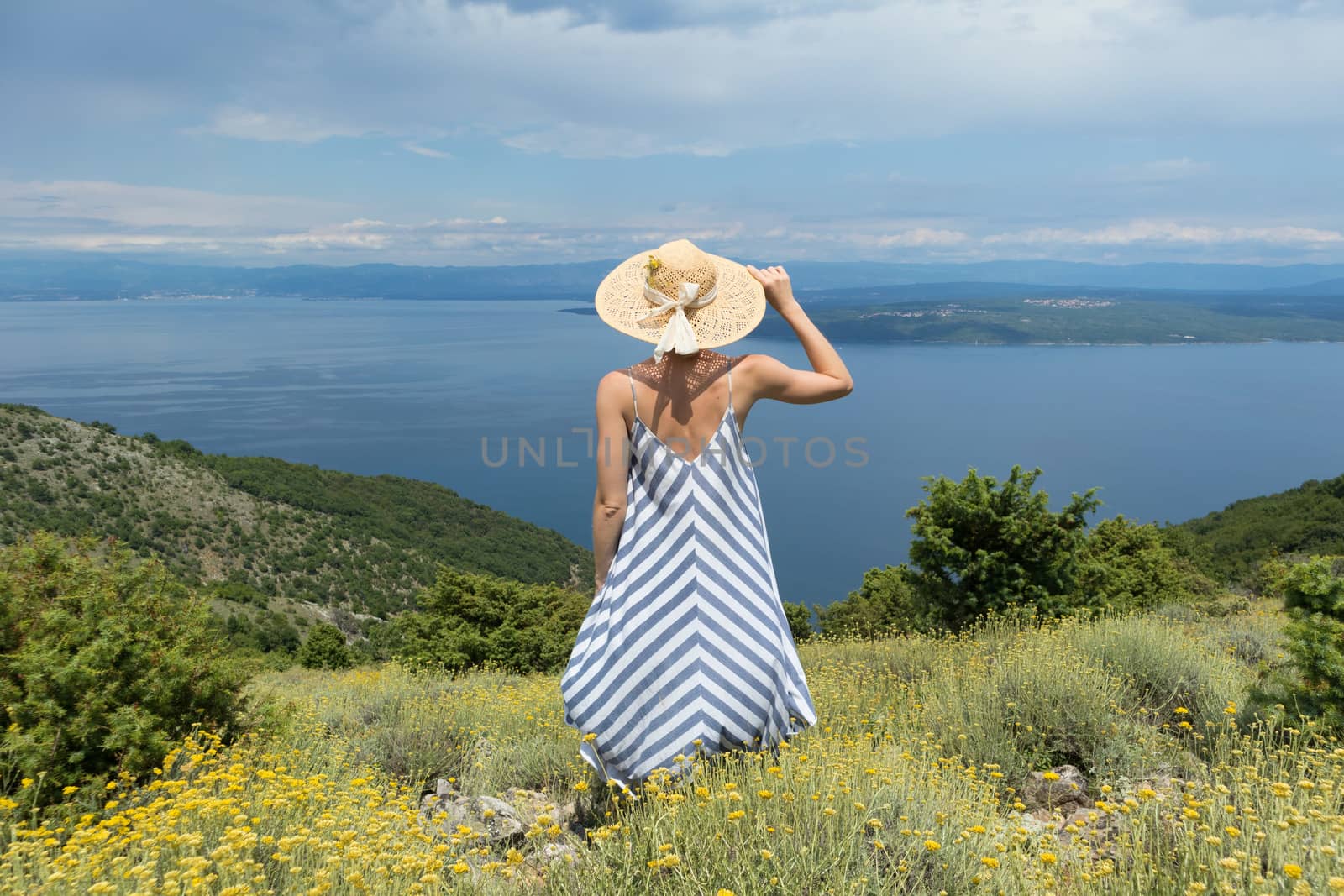 Rear view of young woman wearing striped summer dress and straw hat standing in super bloom of wildflowers, relaxing with hands up to the sky, enjoing beautiful view of Adriatic sea nature, Croatia by kasto