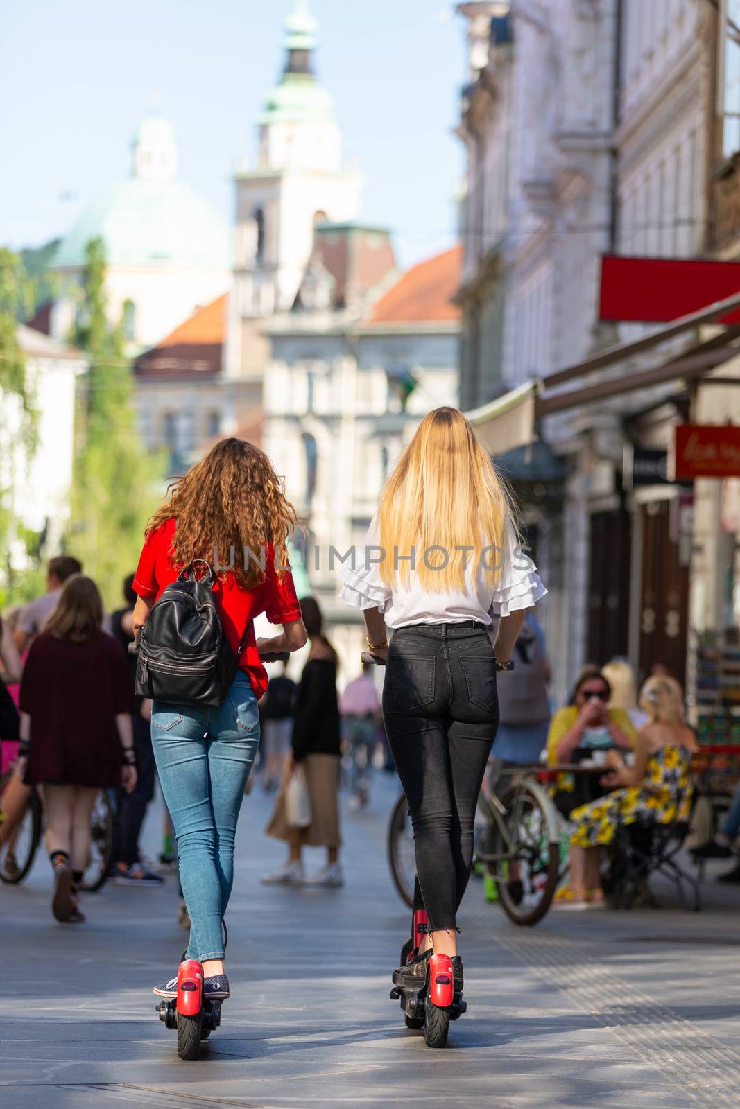 Rear view of trendy fashinable teenager girls riding public rental electric scooters in urban city environment. New eco-friendly modern public city transport in Ljubljana, Slovenia.