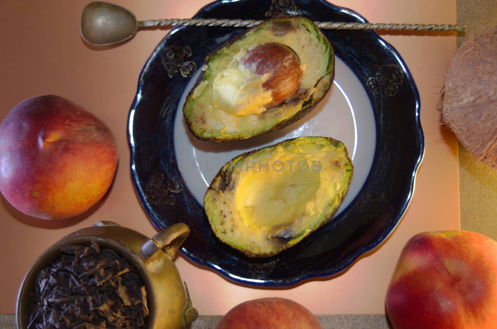food composition - two avocado halves on a plate and a twisted spoon, three ripe peaches, an antique copper cup with large leaf tea and a coconut