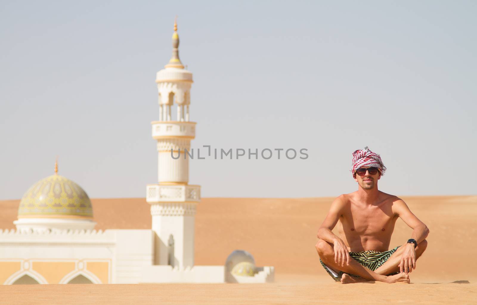 Man traveling in Oman, topless wearing a turban and shorts, sitting on send dune in fron of mosque in desert Oman.