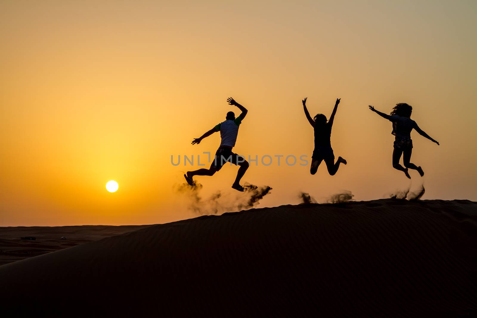 Silhouette of happy traveling people jumping on sand dune and throwing sand in the air in golden sunset hour.