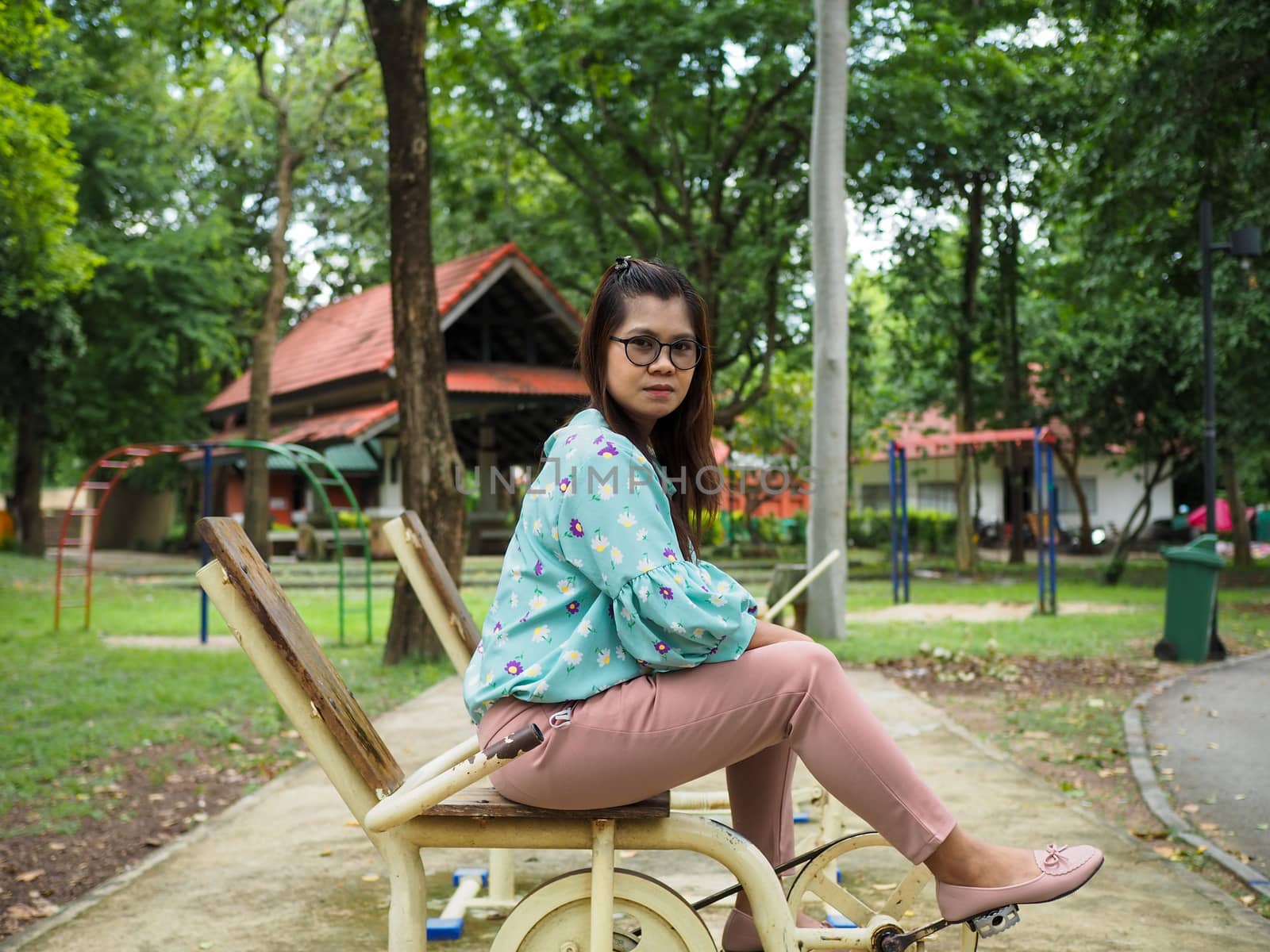 Portrait of an Asian woman Sitting on a gym in the park wearing a green striped shirt Light purple pants With a green tree background.