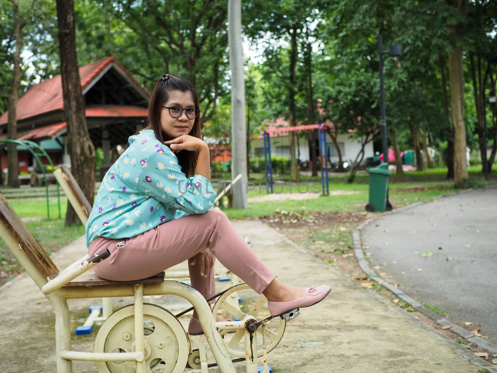 Portrait of an Asian woman Sitting on a gym in the park wearing a green striped shirt Light purple pants With a green tree background.
