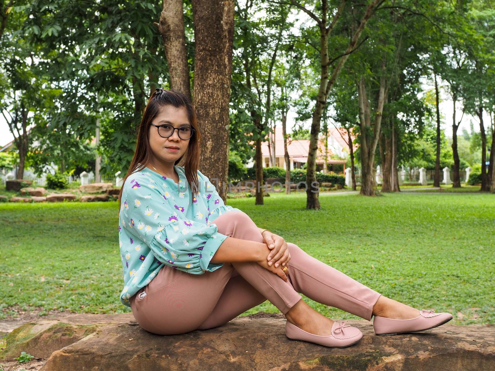 Portrait of an Asiatic woman sitting on a stone wearing a green striped shirt Light purple pants With a green tree background.