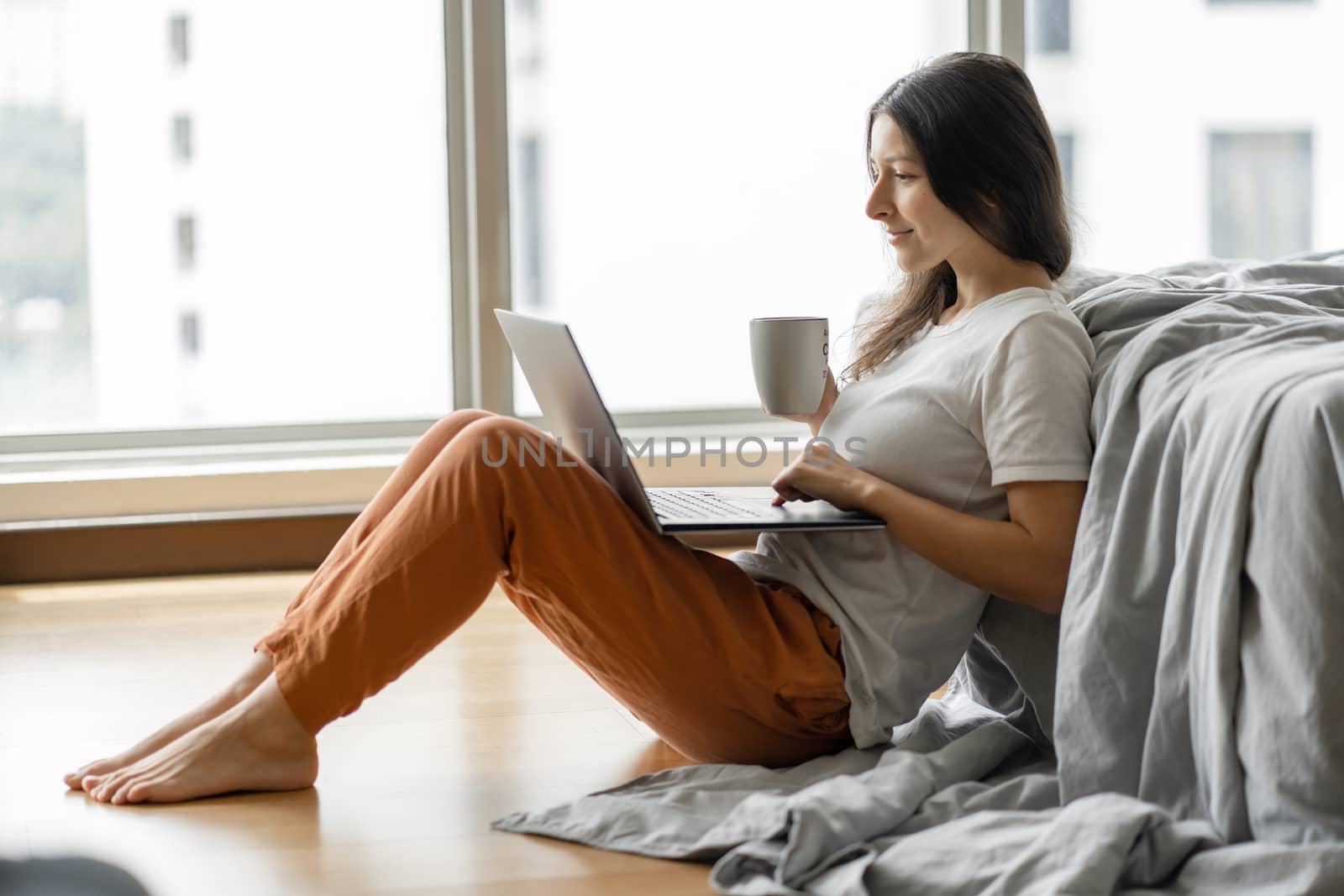 Beautiful young brunette girl working on a laptop and drinking coffee, sitting on the floor near the bed by the panoramic window. Stylish modern interior. A cozy workplace. Shopping on the Internet