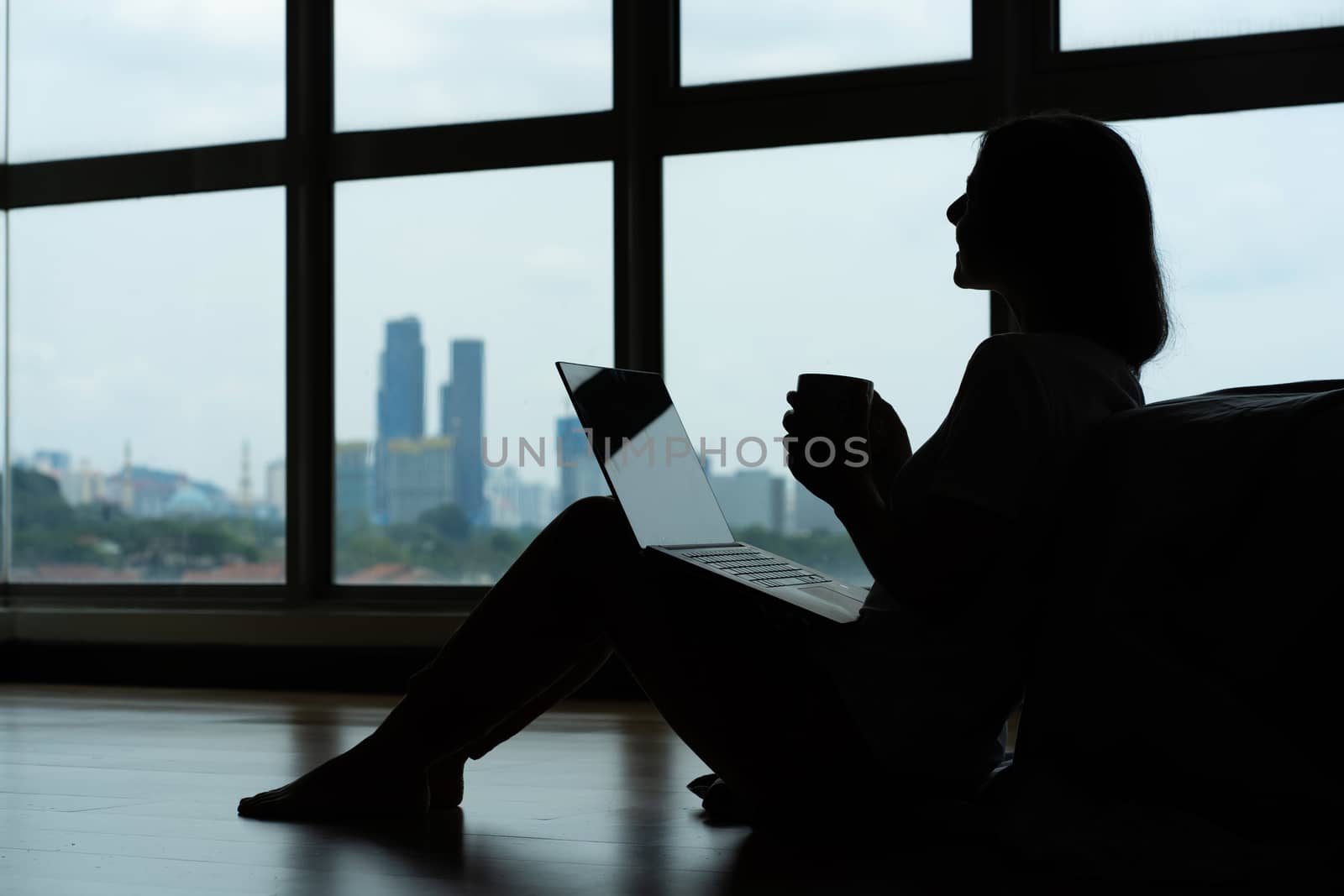 Siluet girl working on a laptop and drinking coffee, sitting on the floor near the bed by the panoramic window. Stylish modern interior. A cozy workplace. Shopping on the Internet. by Try_my_best