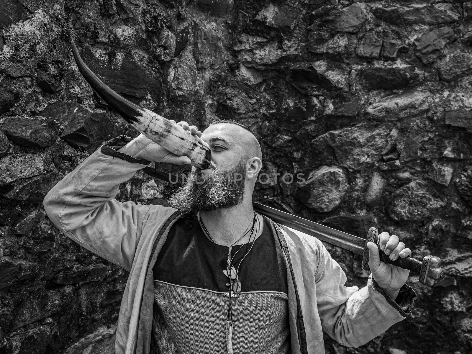 Viking warrior with thick beard and big sword drinks from the horn in front of a stone wall, black and white historical reenactment image