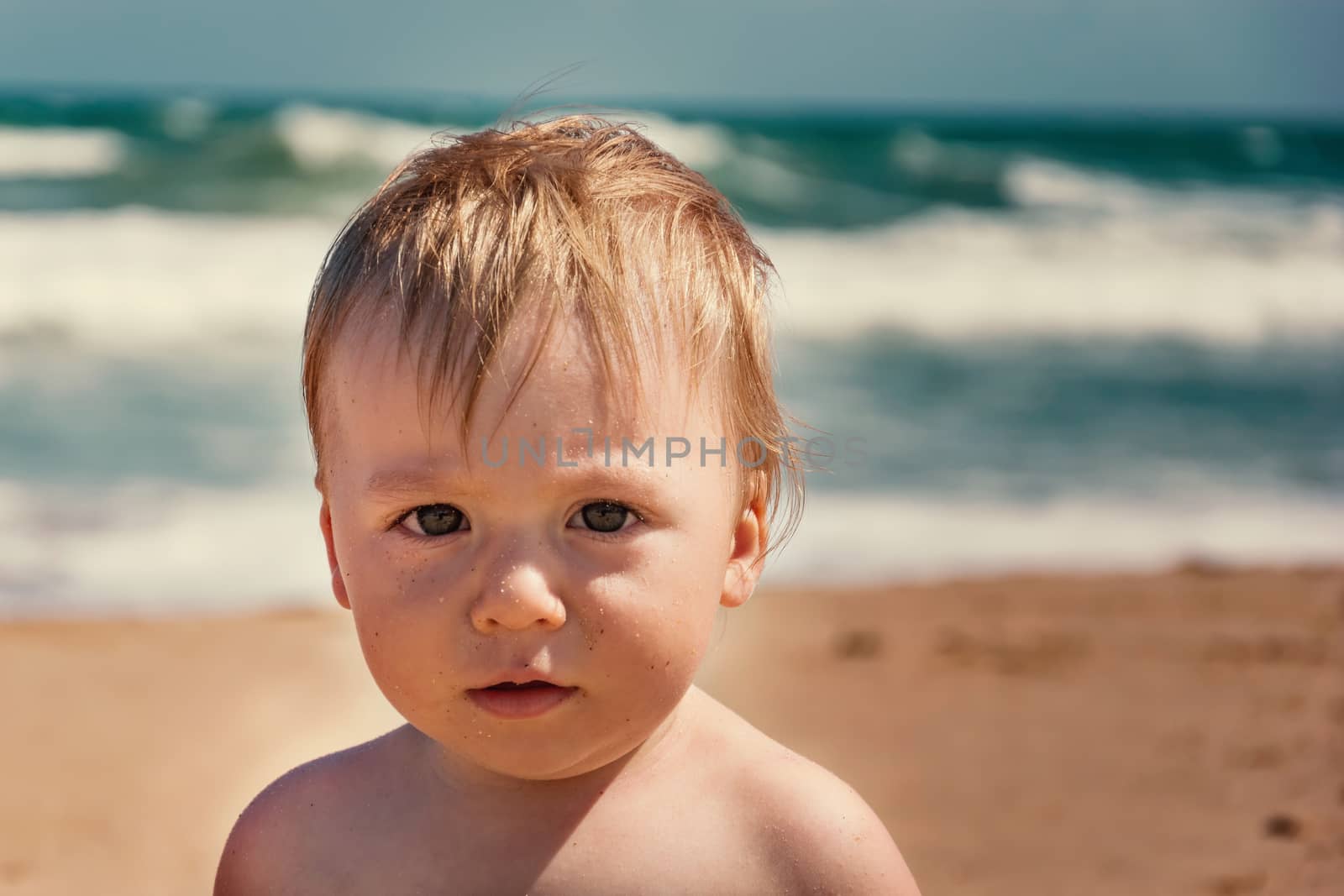 Adorable infant boy looking at camera at summertime. Young caucasian baby at tropical beach with ocean wave behind him. Male kid portrait on his first vacations at sandy beach. Curiosity expression.