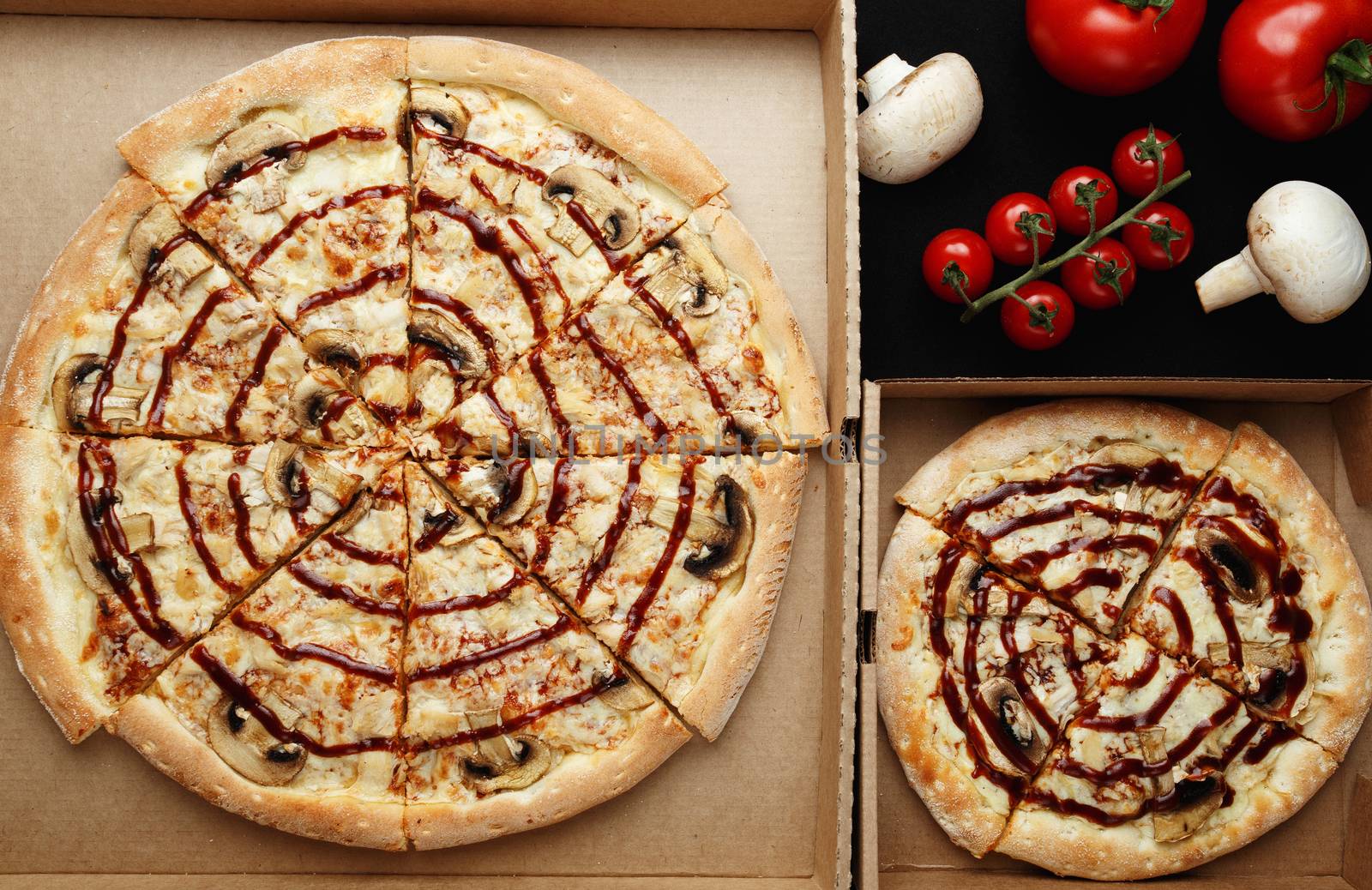 Large and small pizzas lie on an open cardboard box. Next to them are cherry tomatoes and mushroom mushrooms. top view photo
