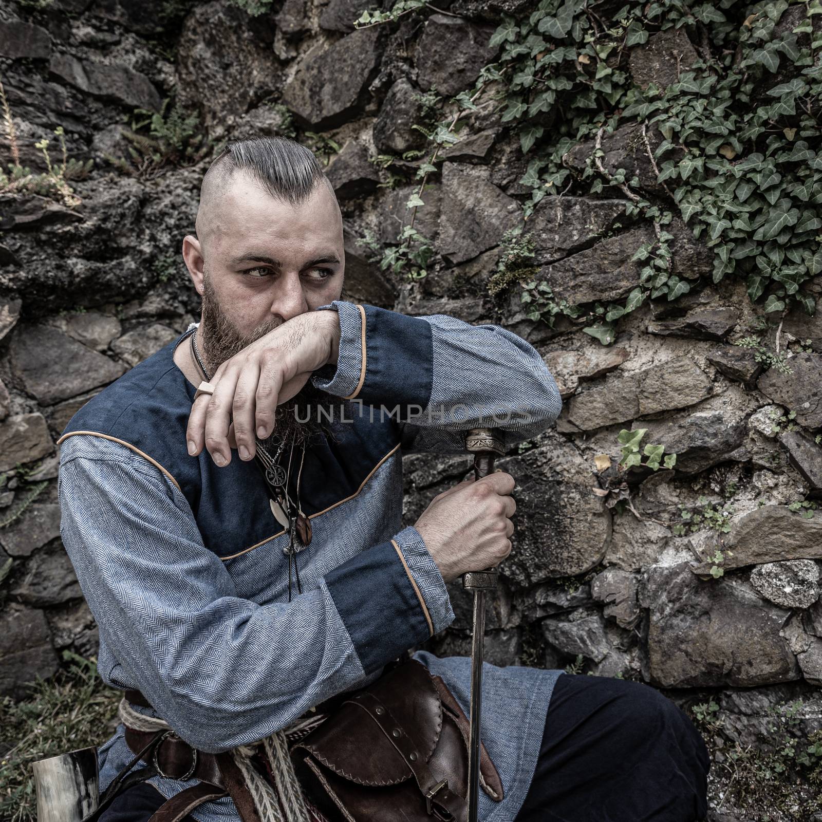 Viking warrior with thick beard sits thoughtfully in front of a stone wall, image of historical re-enactment among medieval ruins