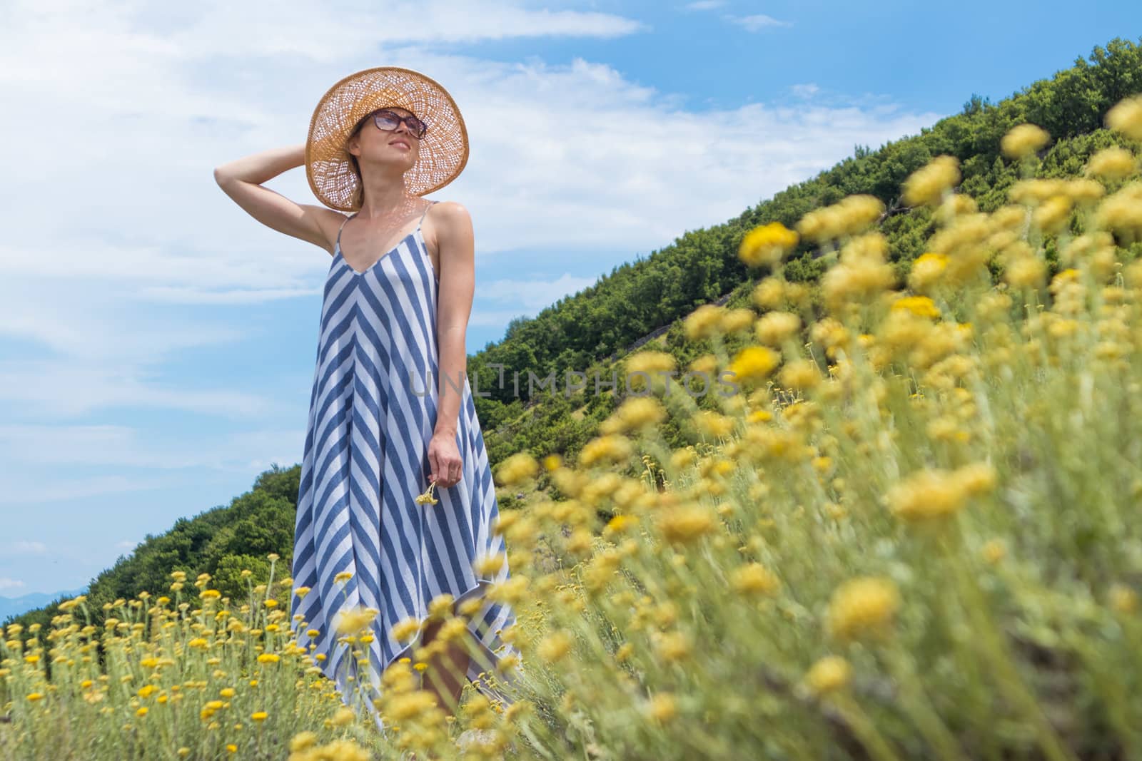 Young woman wearing striped summer dress and straw hat standing in super bloom of wildflowers, relaxing while enjoing beautiful nature of of Adriatic sea coastal nature of Croatia. by kasto