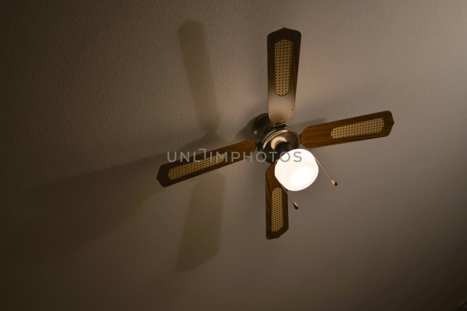 Mediterranean brown wood and material ceiling fan casts a long shadow on a plaster ceiling.