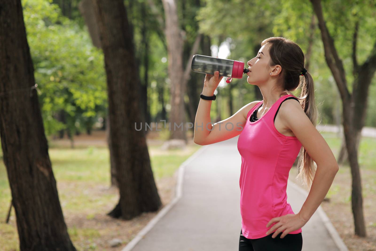 Beautiful fitness athlete runner woman drinking water in the park. Portrait face of a young woman holding a water bottle in summer. Forest