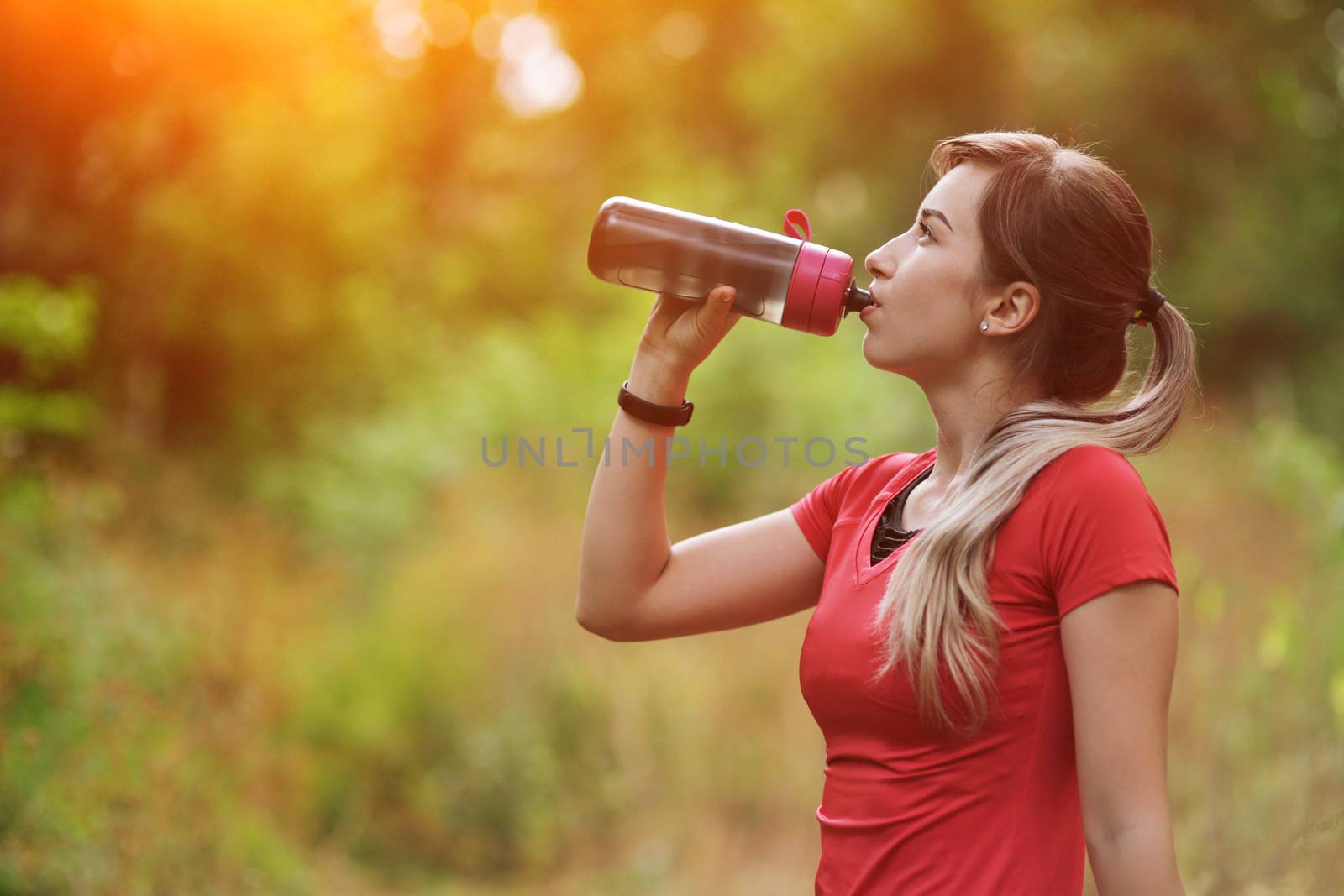 Beautiful fitness athlete runner woman drinking water in the park. Portrait face of a young woman holding a water bottle in summer. Forest