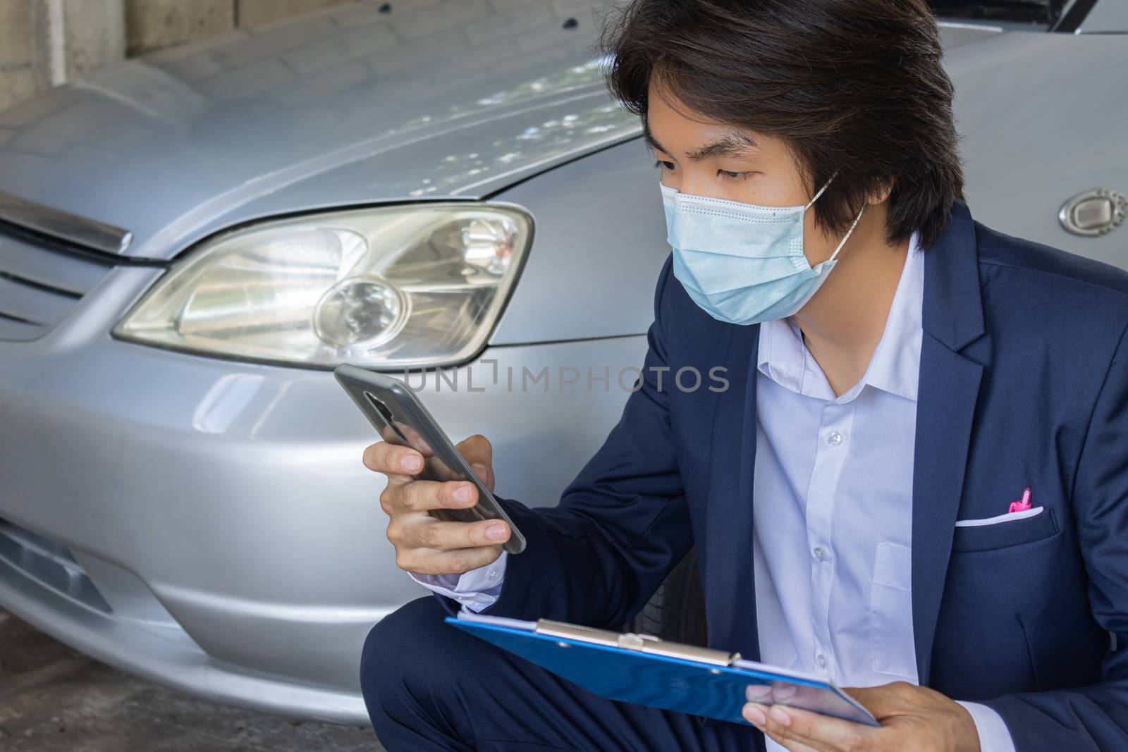 Sitting Asian Insurance Agent or Insurance Agency in Suit Wear Mask and See Smartphone and Hold Report Inspecting Car from Accident for Claim at Garage