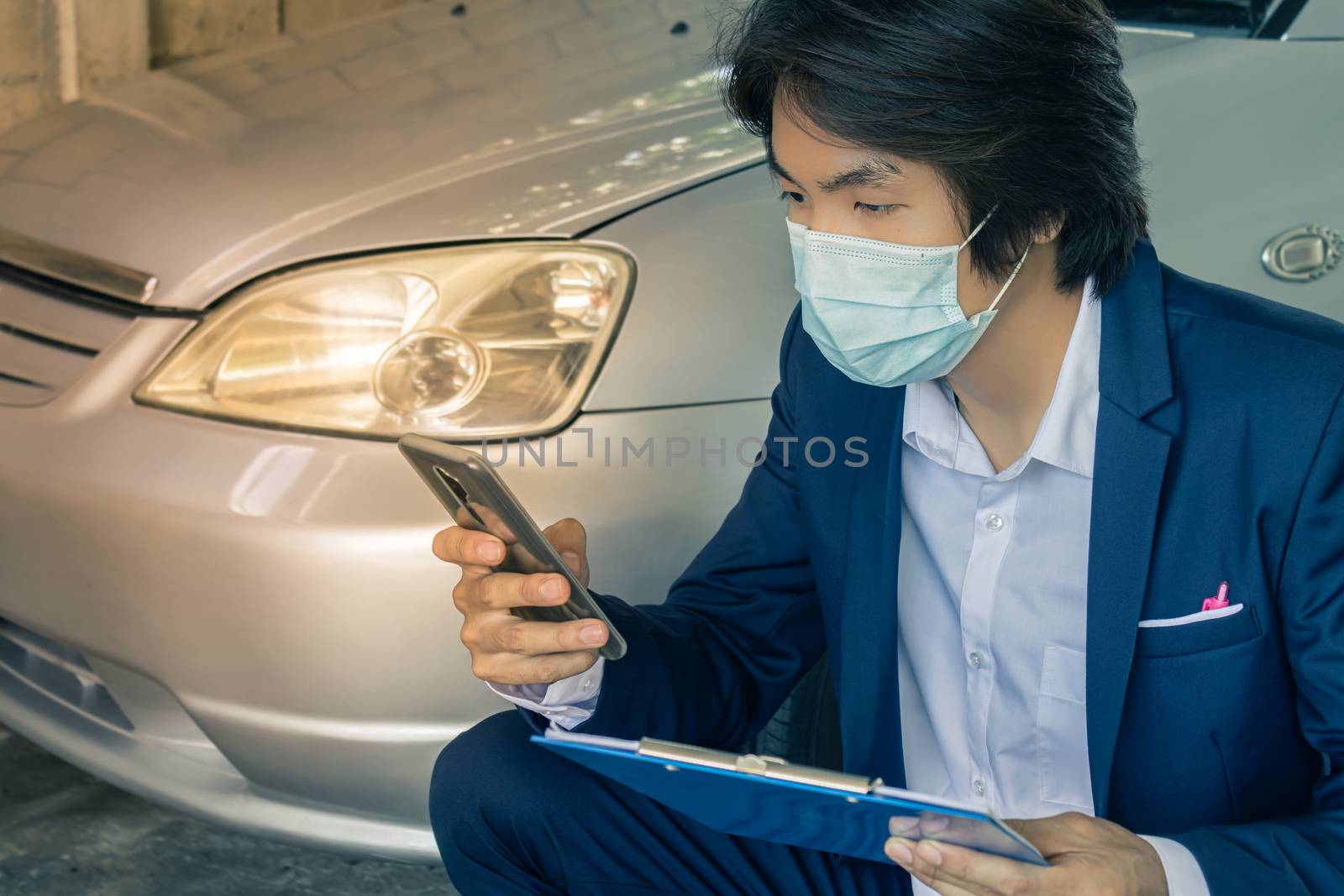 Sitting Asian Insurance Agent or Insurance Agency in Suit Wear Mask and See Smartphone and Hold Report Inspecting Car from Accident for Claim at Garage in Vintage Tone