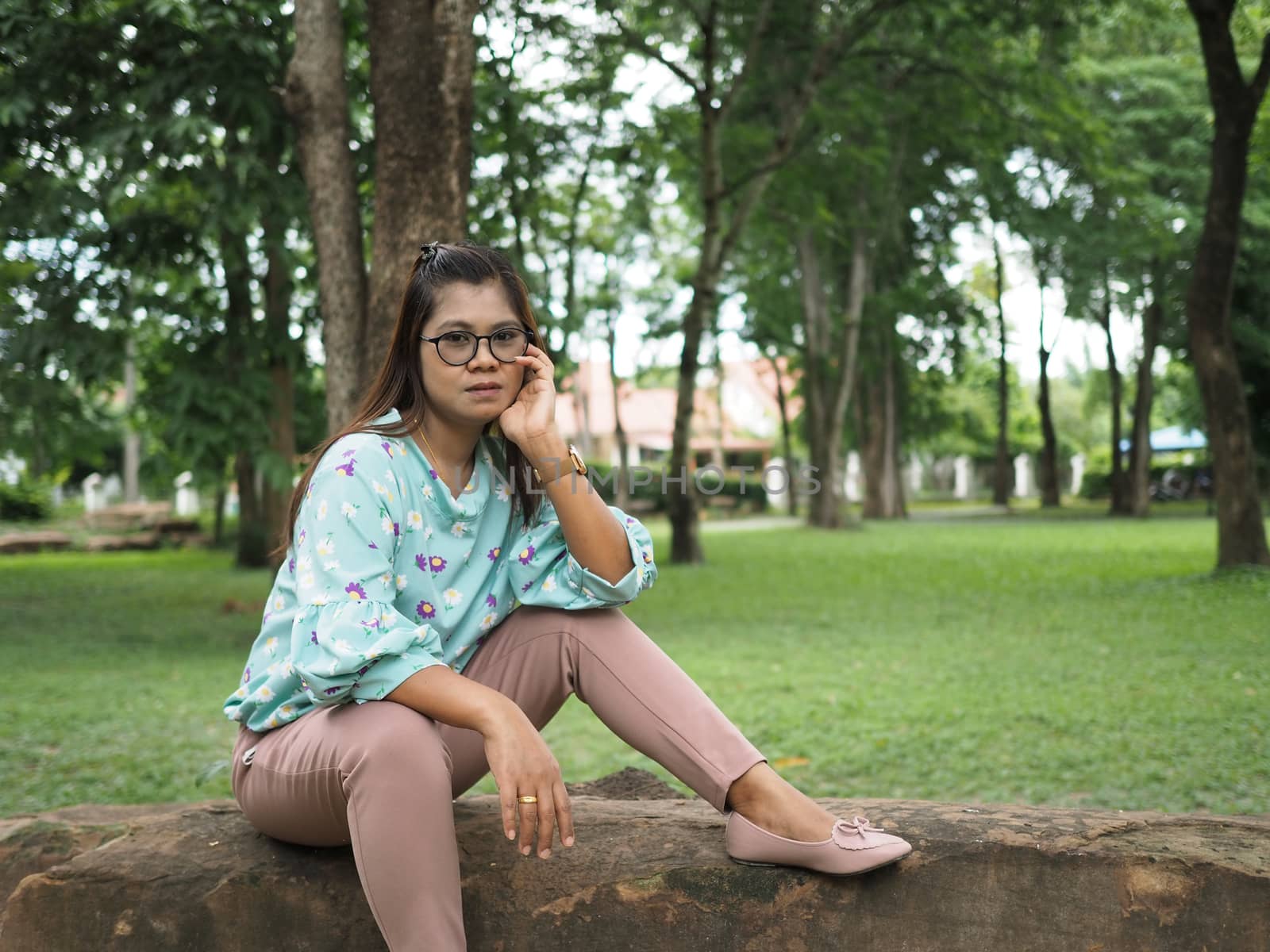 Portrait of an Asian woman With long hair sitting on a stone in a garden with a green tree background.