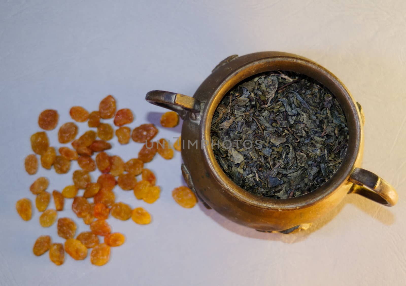 copper vase with large leaf tea next to scattered raisins on a light background