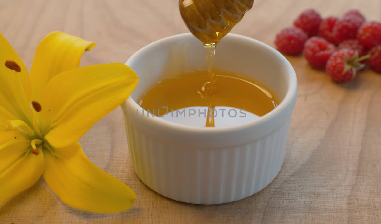 Close up honey pouring from honey spoon into a bowl. Raspberry and lily on a table in the garden at summer day. Healthy fresh food concept.