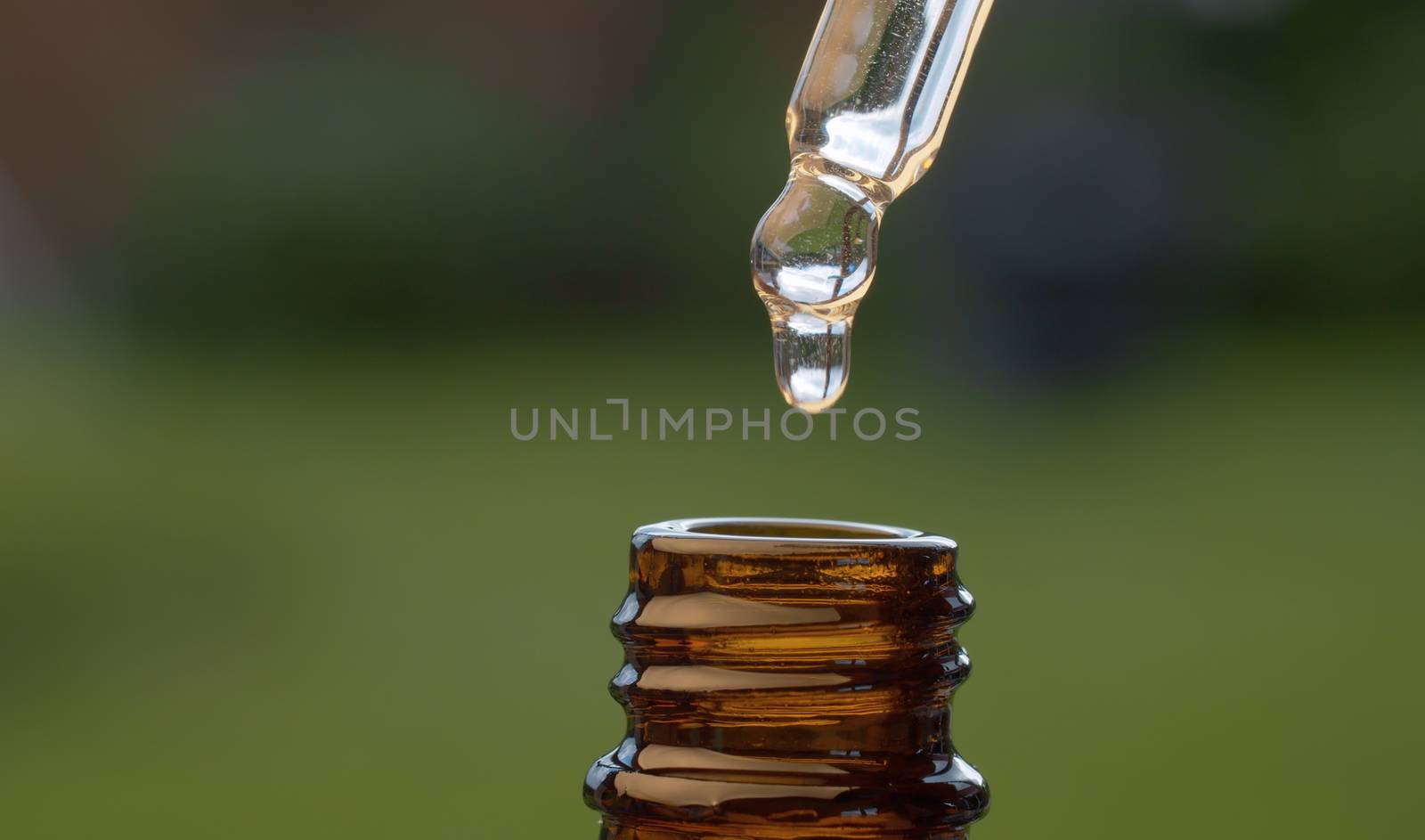 Macro shot essential oil dripping from a pipette on blurry natural background. Extreme close up hand taking transparent oil from a brown glass bottle