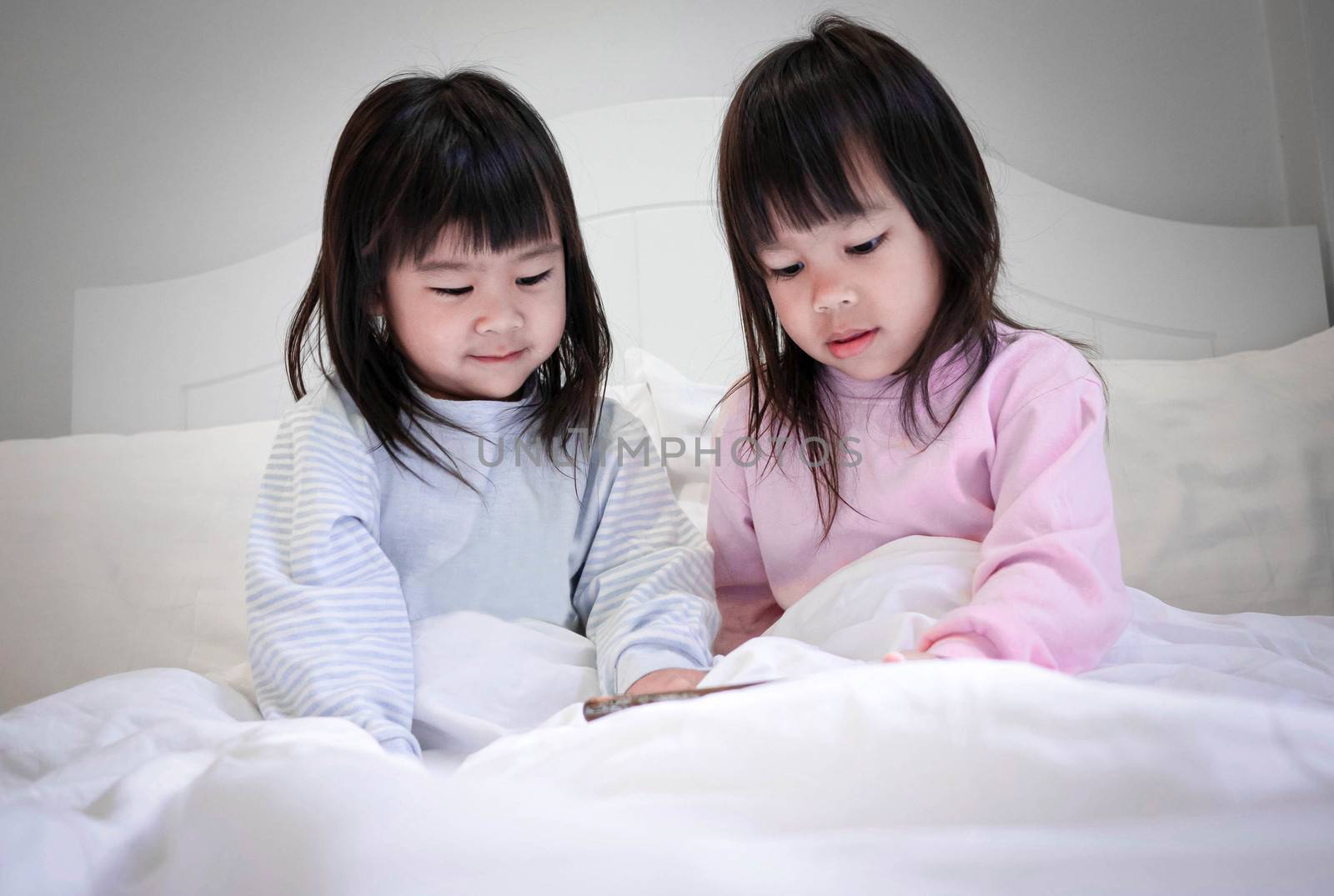 Asian sibling girls in pajamas watching smartphone on a bed at n by TEERASAK