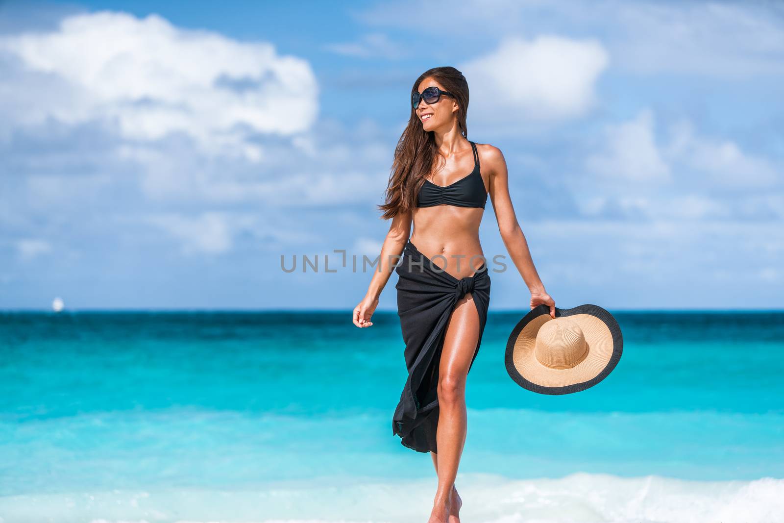 Beach vacation luxury travel elegant bikini woman in fashion swimwear sun tanning in tropical destination getaway. Asian girl wearing cover-up pareo, sunglasses, hat. Body skin care suntan protection by Maridav