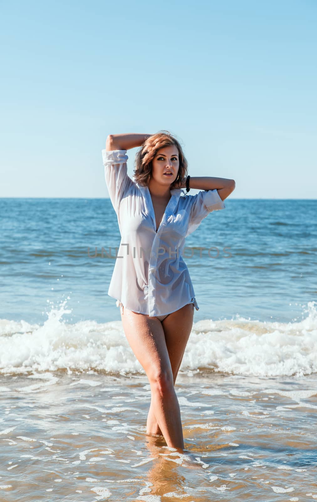 young blonde woman in a wet white shirt standing in the water near the seashore with her hands behind her head on sunny summtr day